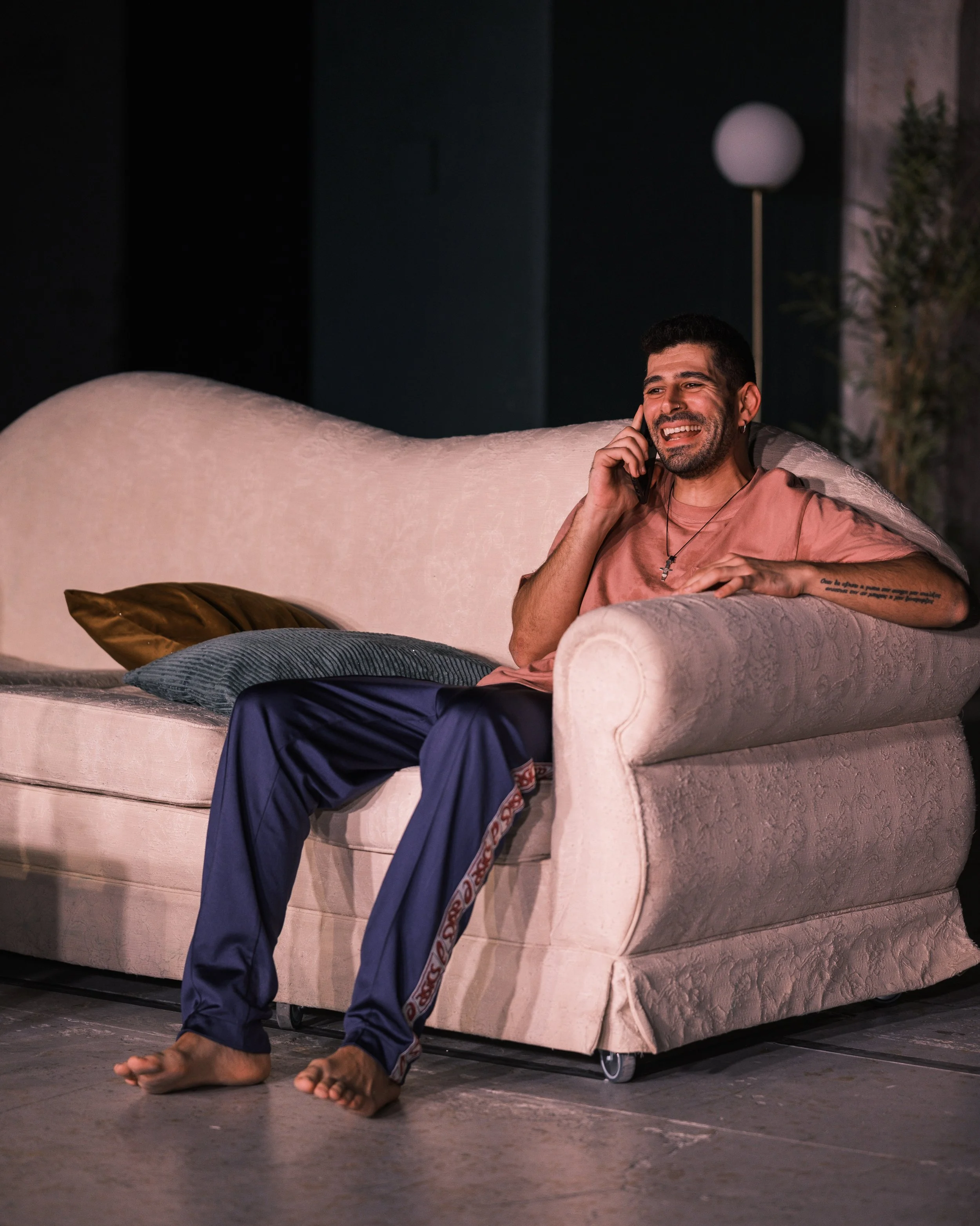 A man with short dark hair and a beard sitting on a beige sofa, smiling and talking on a mobile phone. He is wearing a pink T-shirt and navy pajama pants with a decorative stripe along the side. There are two pillows on the sofa, one brown and one gr