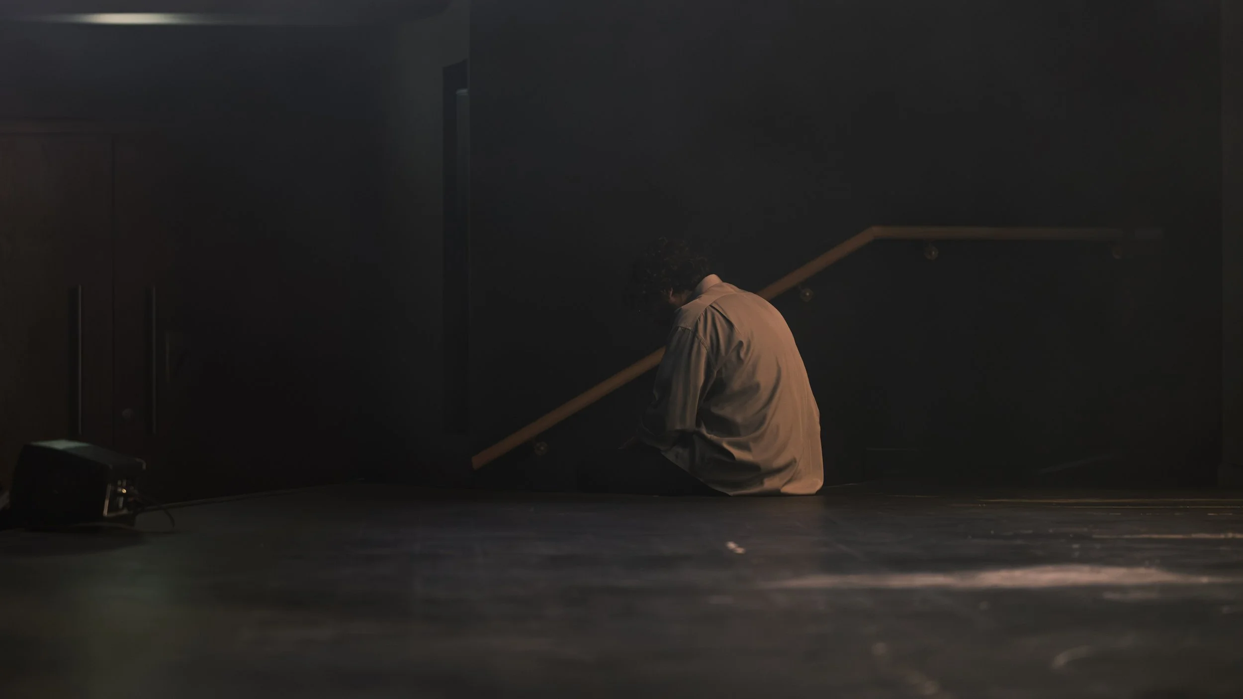A person with curly hair in a beige shirt sitting on the floor with their head bowed, facing a dark wall with stairs and a handrail.