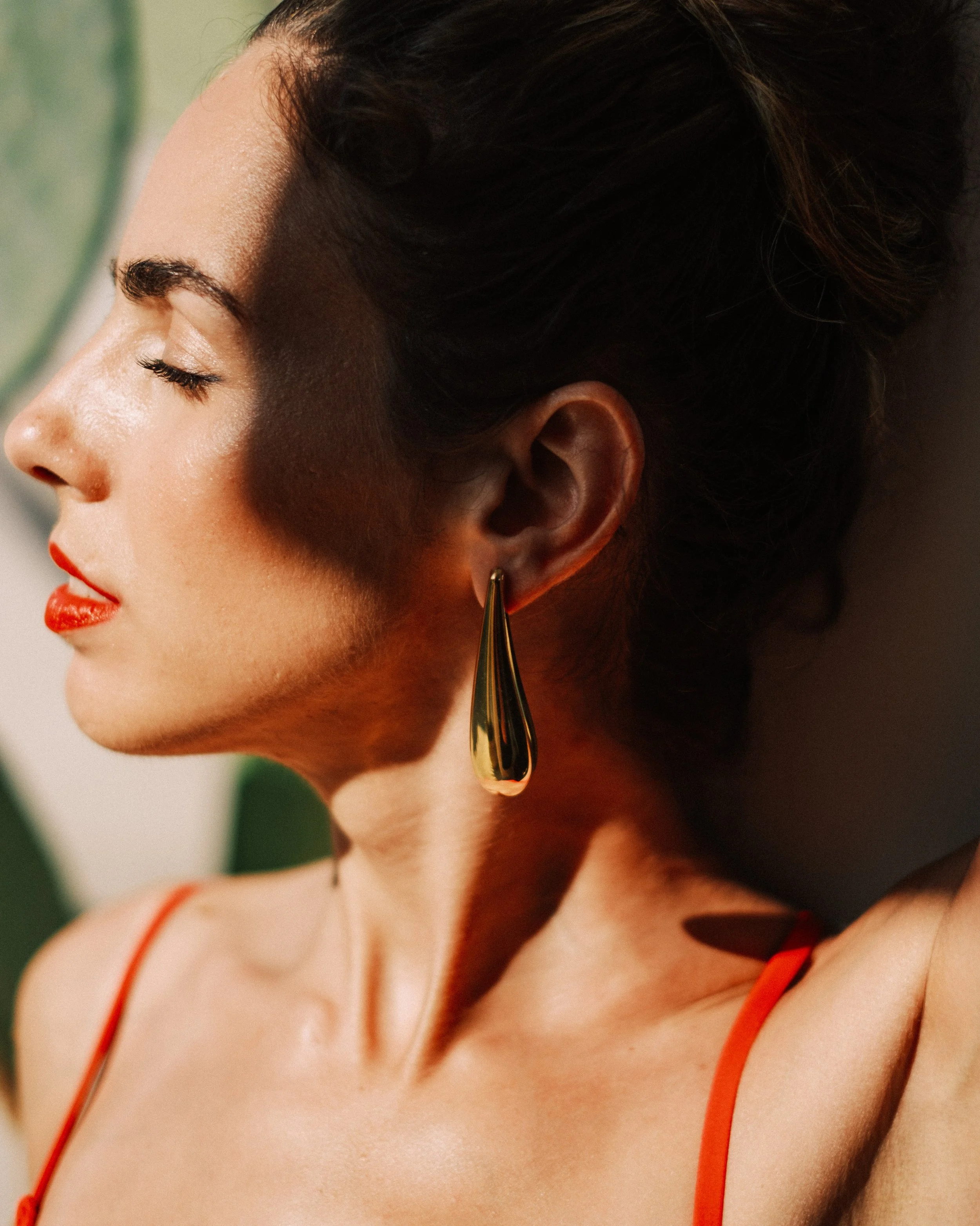 Close-up of a woman with closed eyes, wearing bright red lipstick, and a large gold teardrop-shaped earring, with dark hair styled up, in a warm lighting setting.