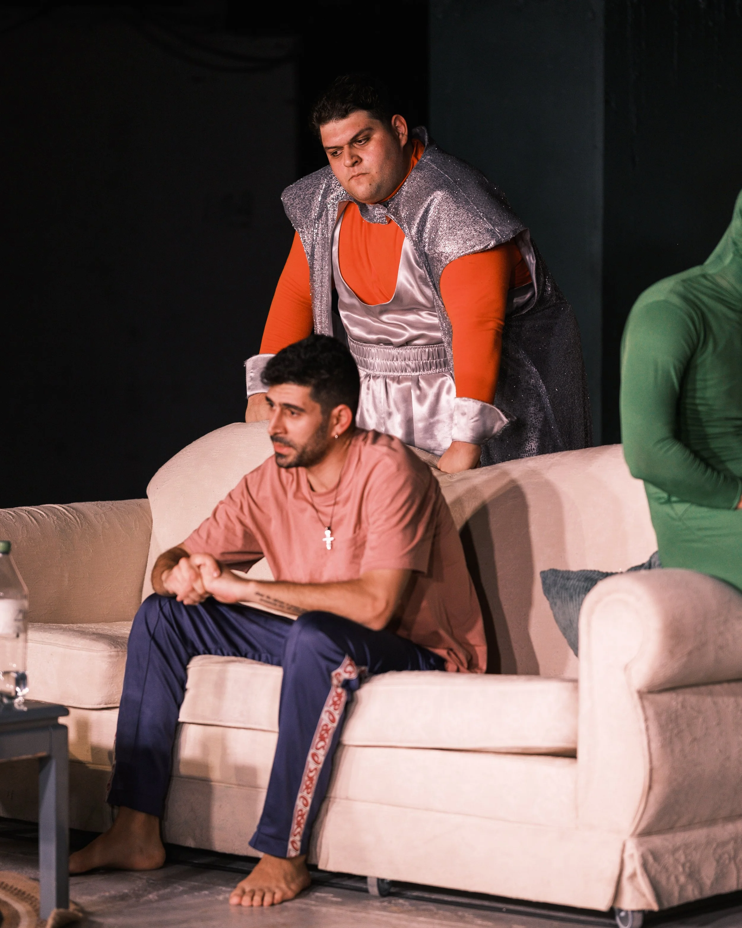 Two men sitting on a beige couch, one in a pink shirt and blue sweatpants with a cross necklace, the other behind him in a shiny, colorful costume, on a stage with dark background.