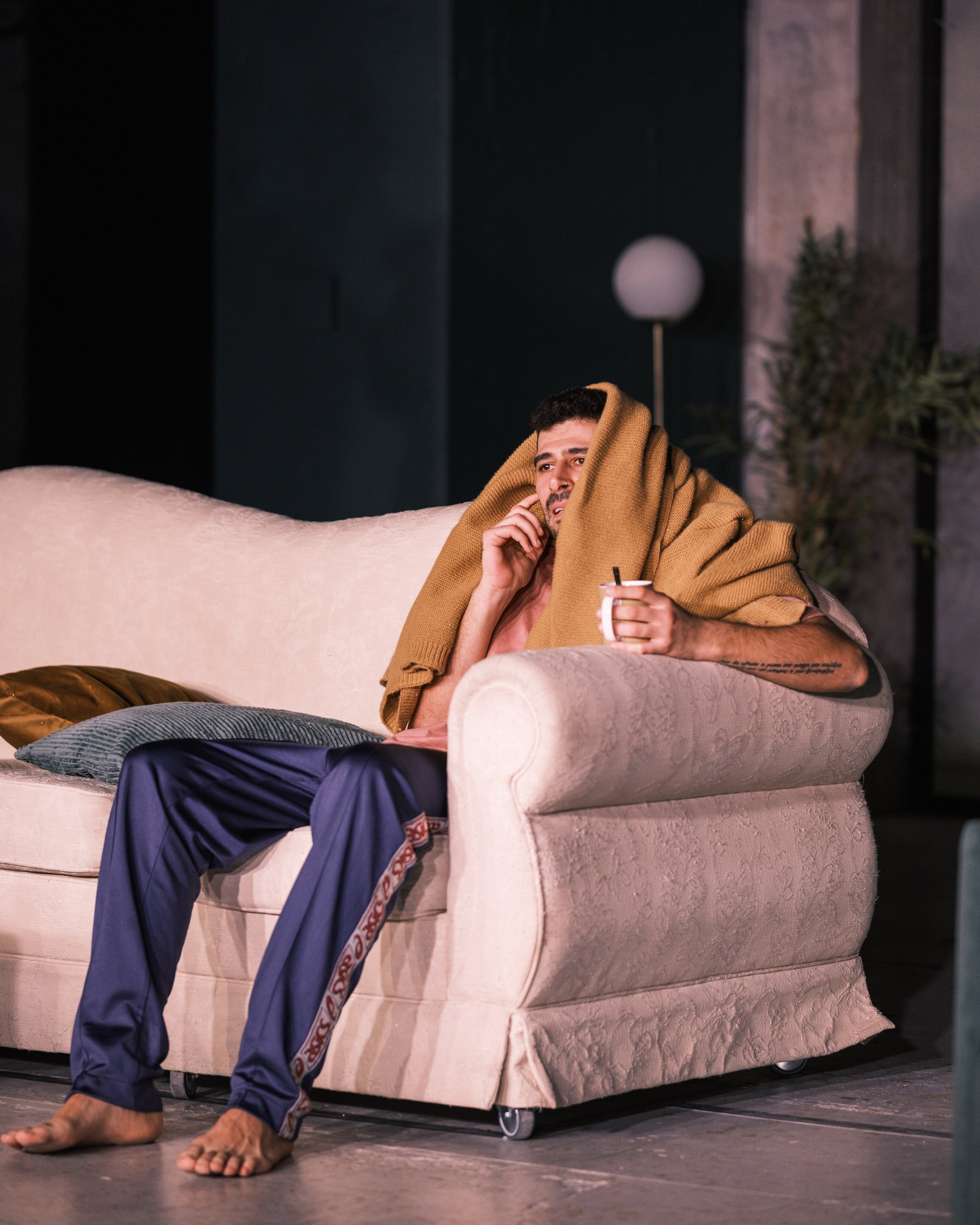 A man sitting on a beige couch, holding a mug with a straw, wrapped in a brown blanket, looking worried or pensive, in a dimly lit room with a lamp and some plants in the background.
