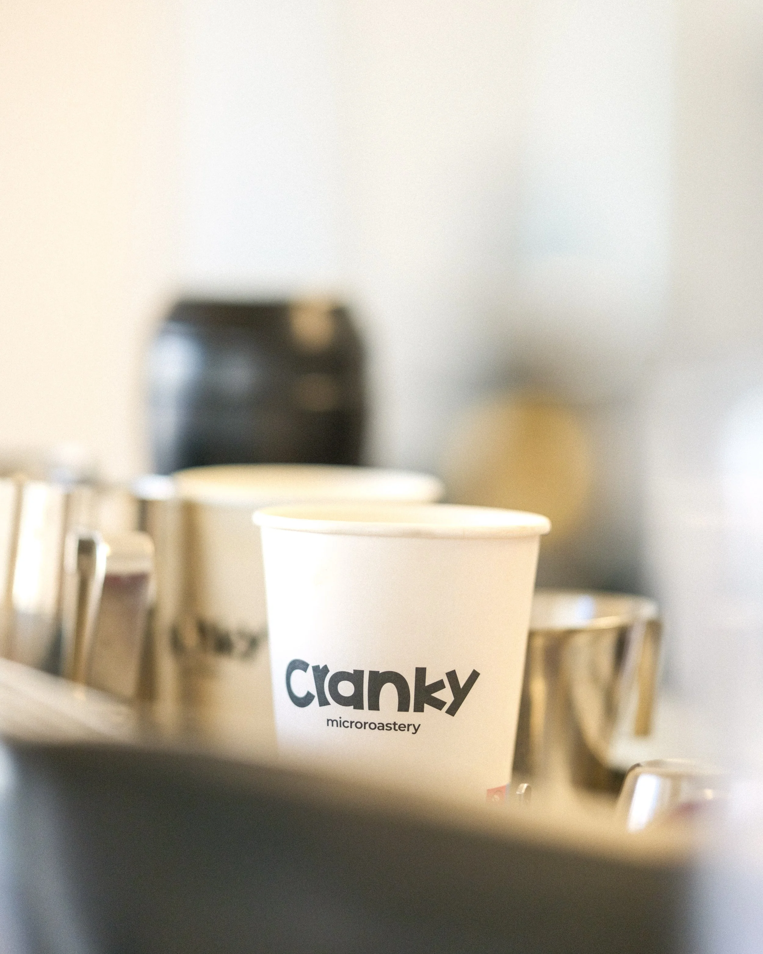 A white paper cup with the logo 'clanky' and the word 'microroastery' printed on it, surrounded by metallic cups and a black container in the background.