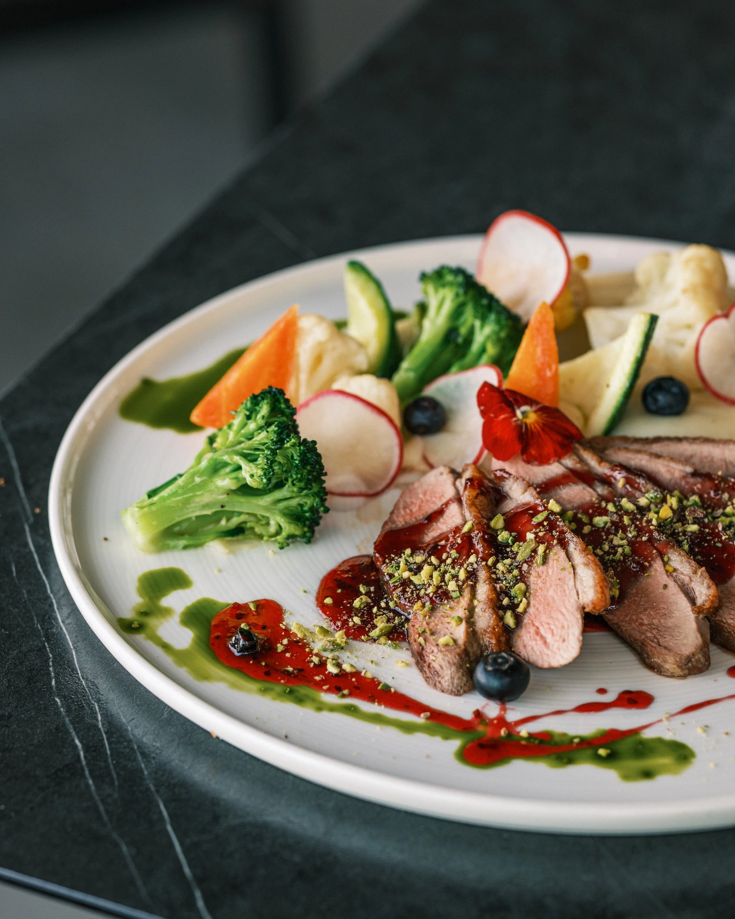 Sliced duck breast with cherry sauce, garnished with chopped pistachios, served with broccoli, radish slices, carrots, cucumber, cauliflower, and blueberries on a white plate.