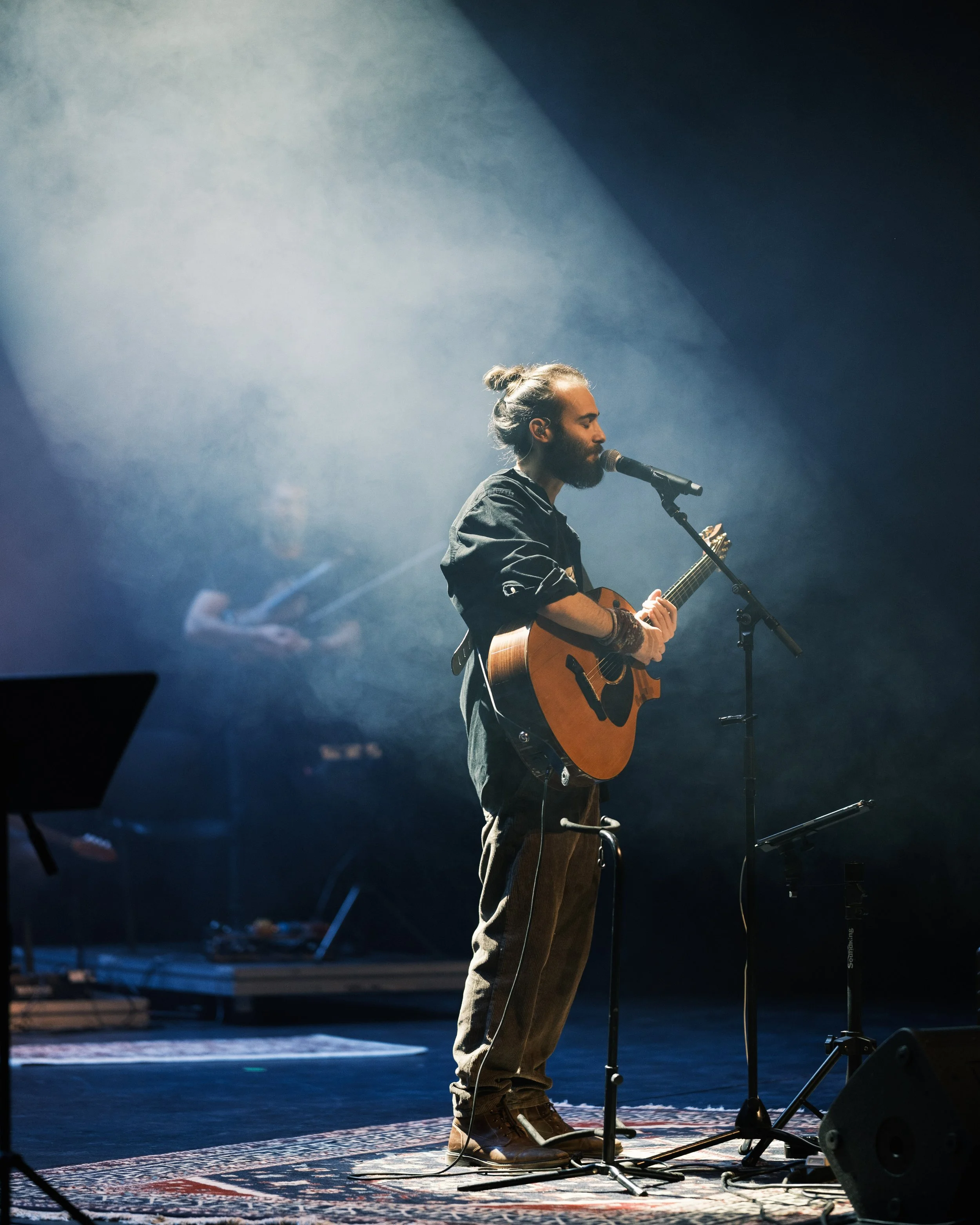 A musician with a beard and hair in a bun playing an acoustic guitar on stage, singing into a microphone, with fog and stage lighting.