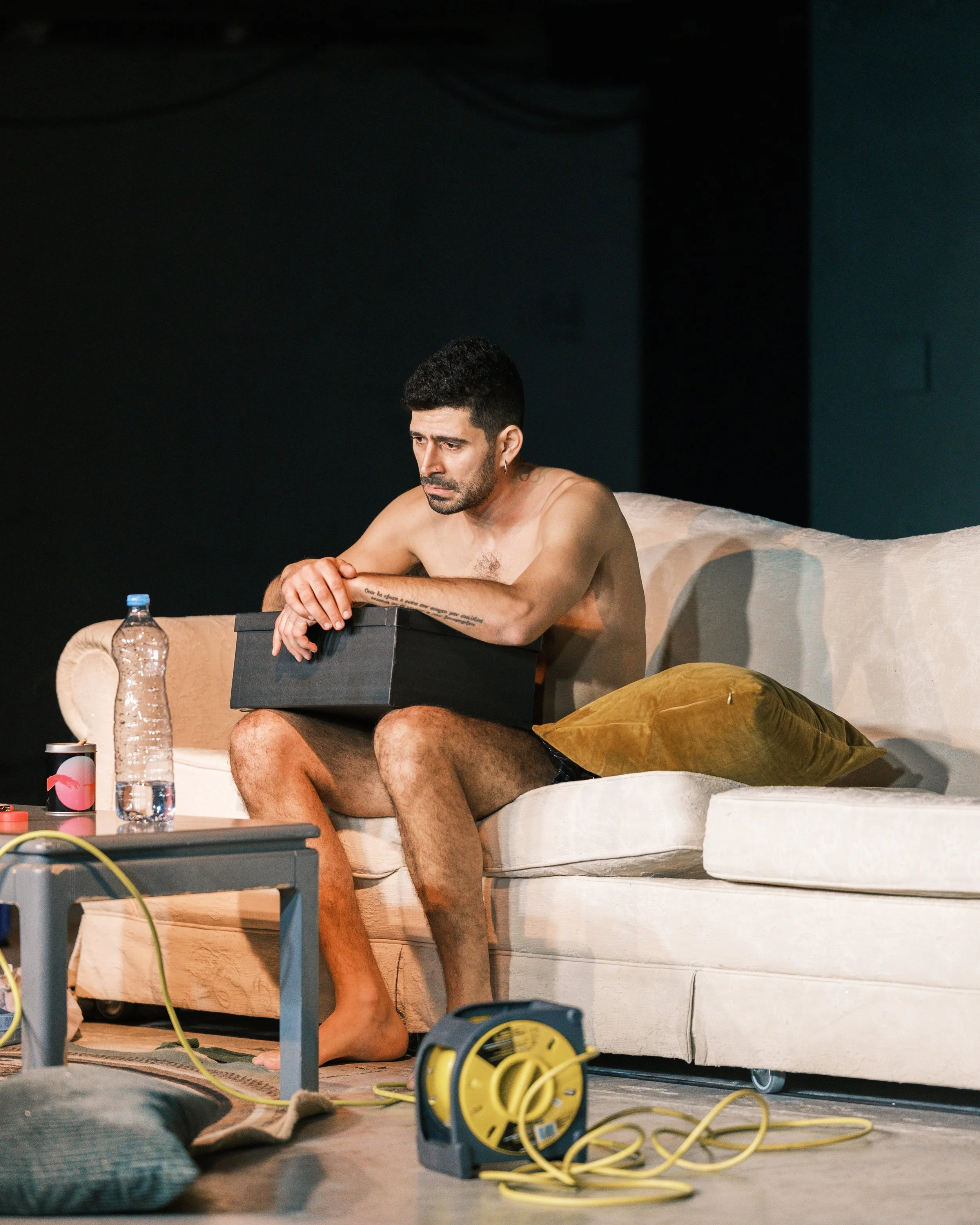 A shirtless man sitting on a beige couch, looking down with a contemplative expression, holding a black box on his lap. There is a yellow extension cord on the floor, a water bottle, a coffee cup, and other items nearby.