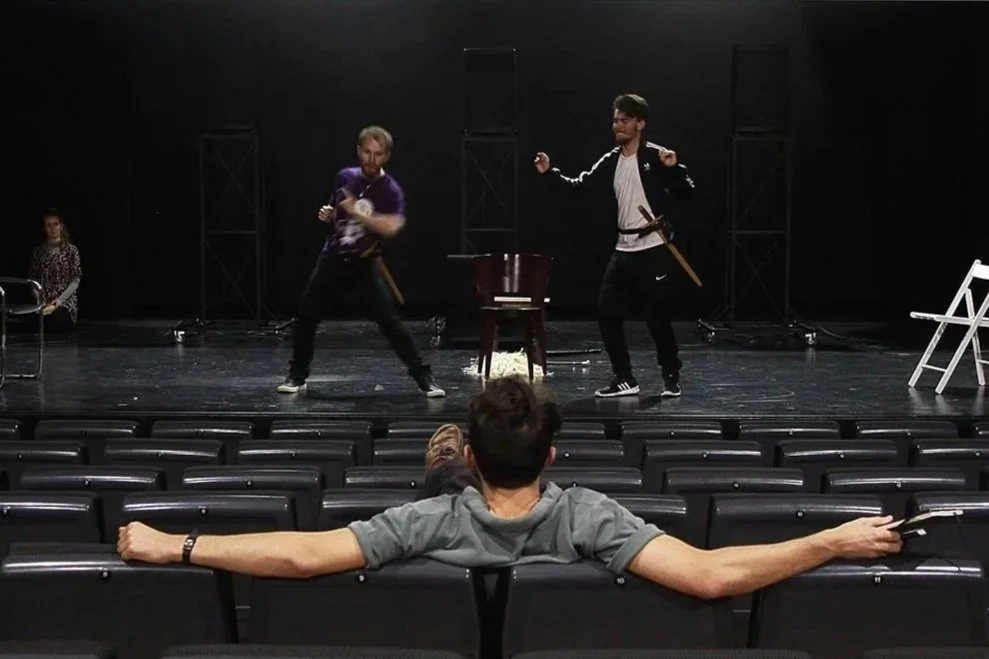 Two men are rehearsing a stage fight with swords, standing in front of a small wooden chair, while a third person watches from the audience with arms outstretched across empty theater seats.