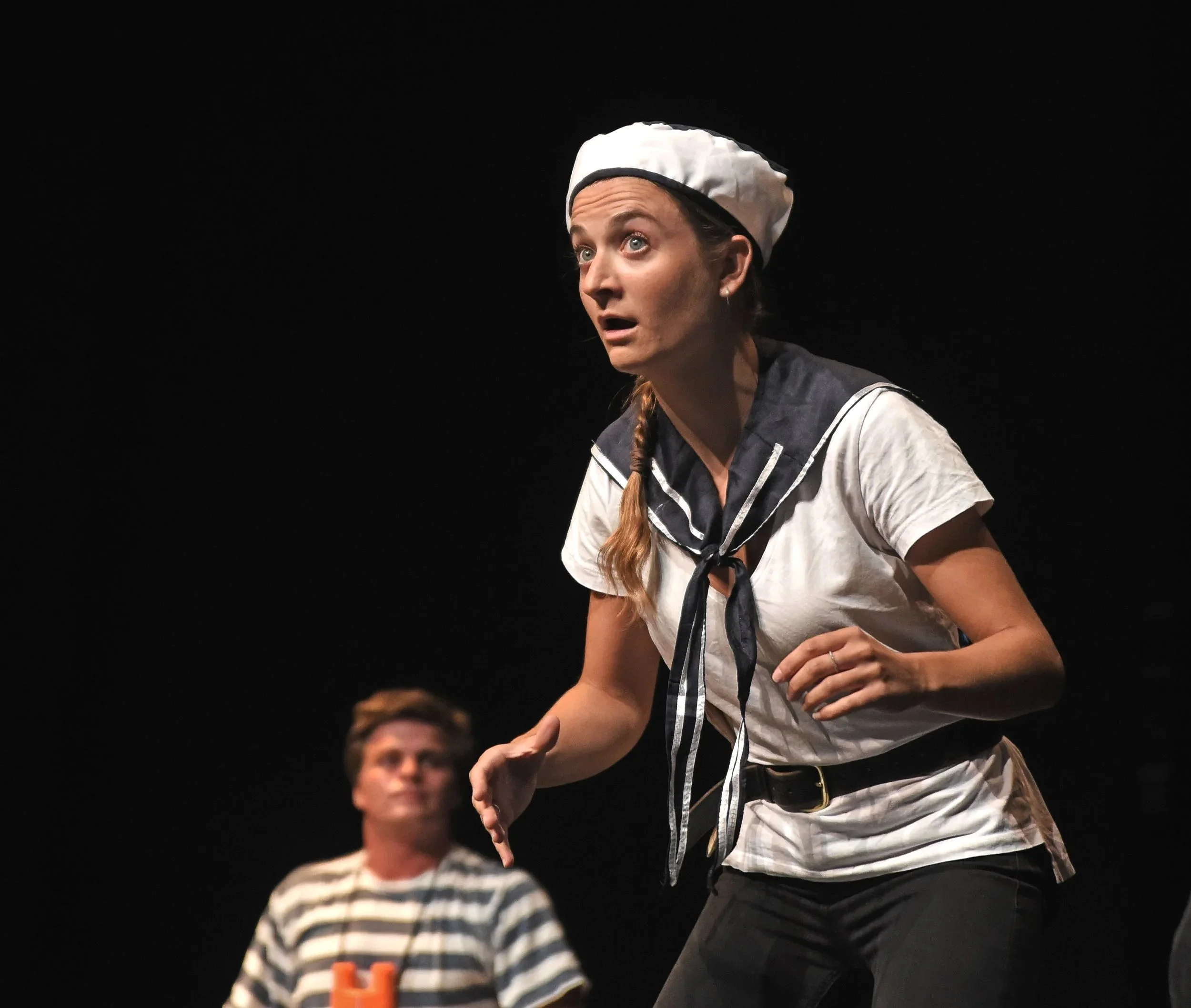 Woman dressed as a sailor with a surprised expression, wearing a white sailor's hat and a striped top, on stage against a black background.