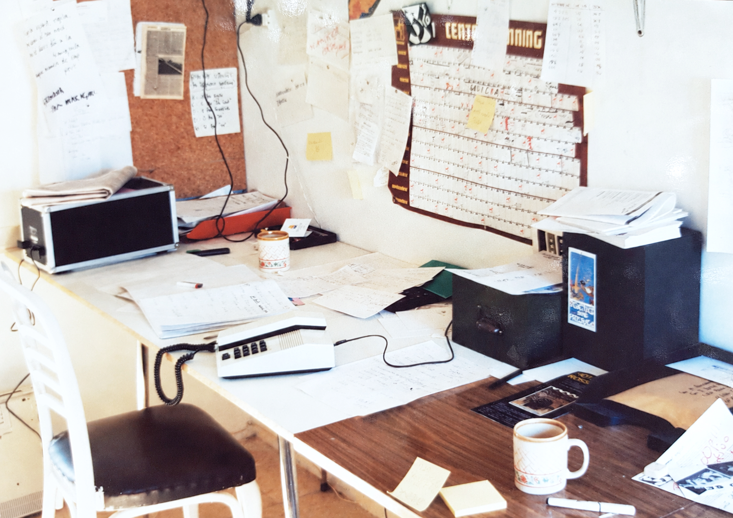 A cluttered desk with papers, books, a coffee mug, and office supplies, with a bulletin board and calendar on the wall.