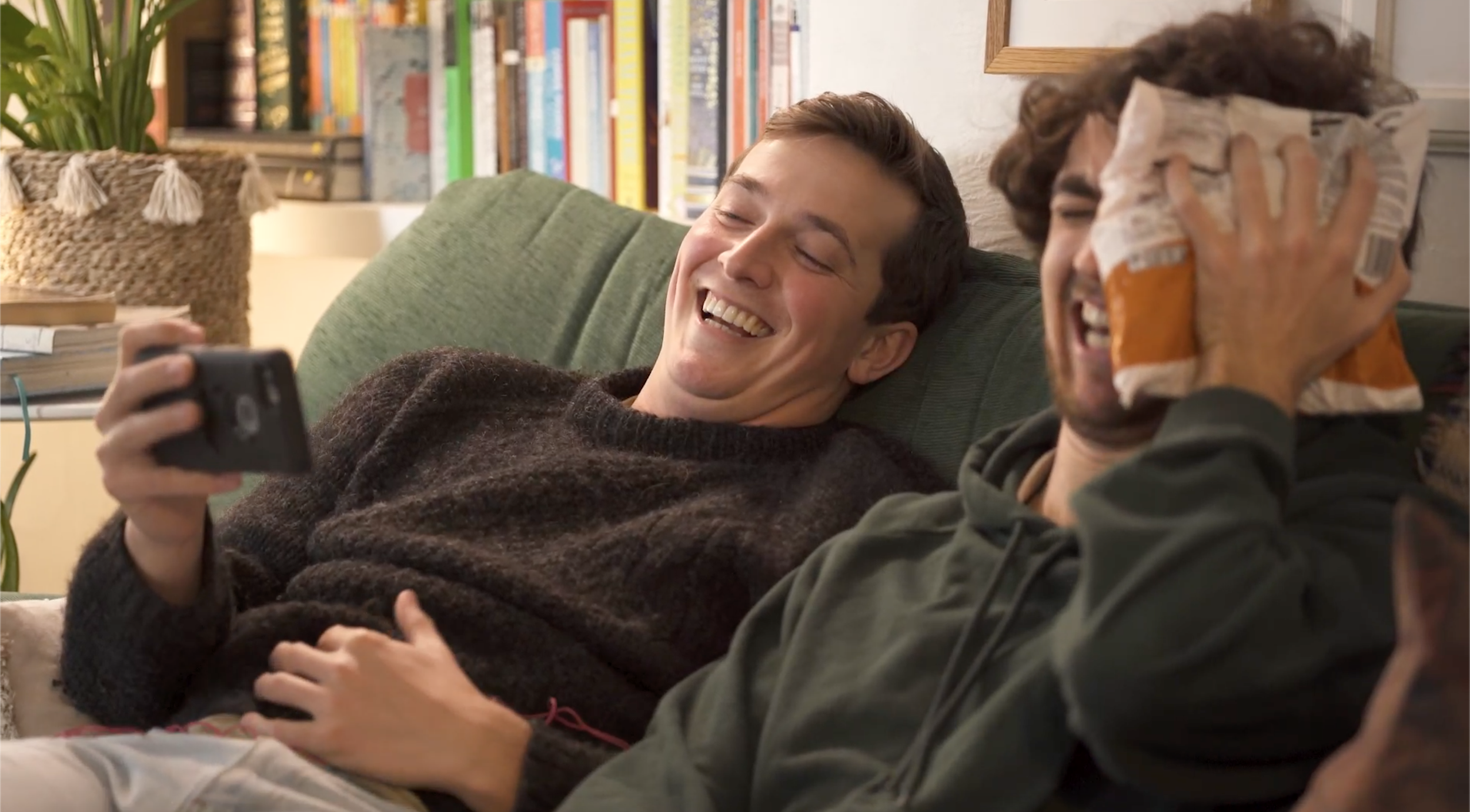 Two young men are sitting on a green couch, smiling and laughing. One is holding a phone, and the other is covering their face with a bag of chips. They are in a cozy living room with books and plants in the background.