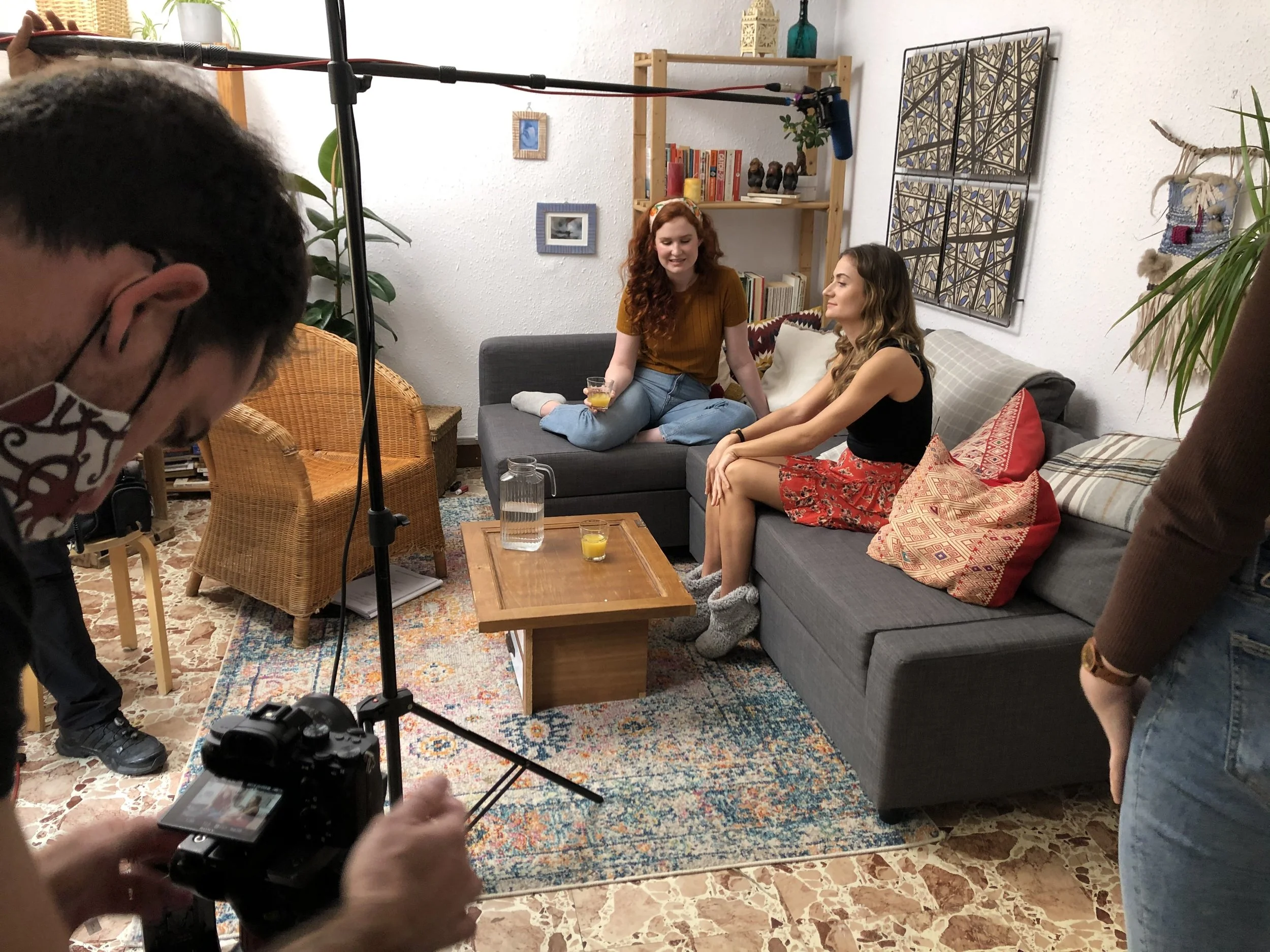 Two women sitting on a gray couch, one with red hair wearing a mustard shirt and jeans, and the other with long brown hair in a black top and red skirt, participating in a filming session with a camera crew recording in a cozy living room. A woman with glasses and a face mask operates the camera, while another person holds a microphone overhead. The room features a colorful patterned rug, a wooden coffee table, a bookshelf, and wall decorations.