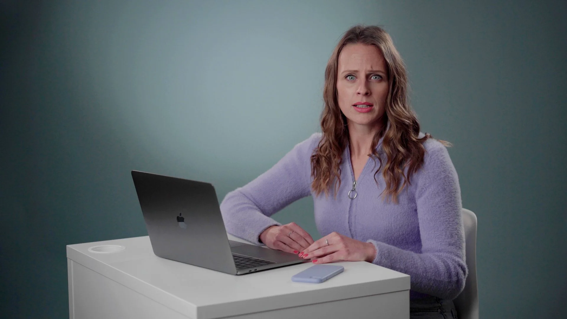 A woman with wavy brown hair and blue eyes sitting at a white desk with an open laptop and a smartphone. She is wearing a light purple zip-up sweater and is looking at the camera with a surprised or confused expression against a plain wall background.