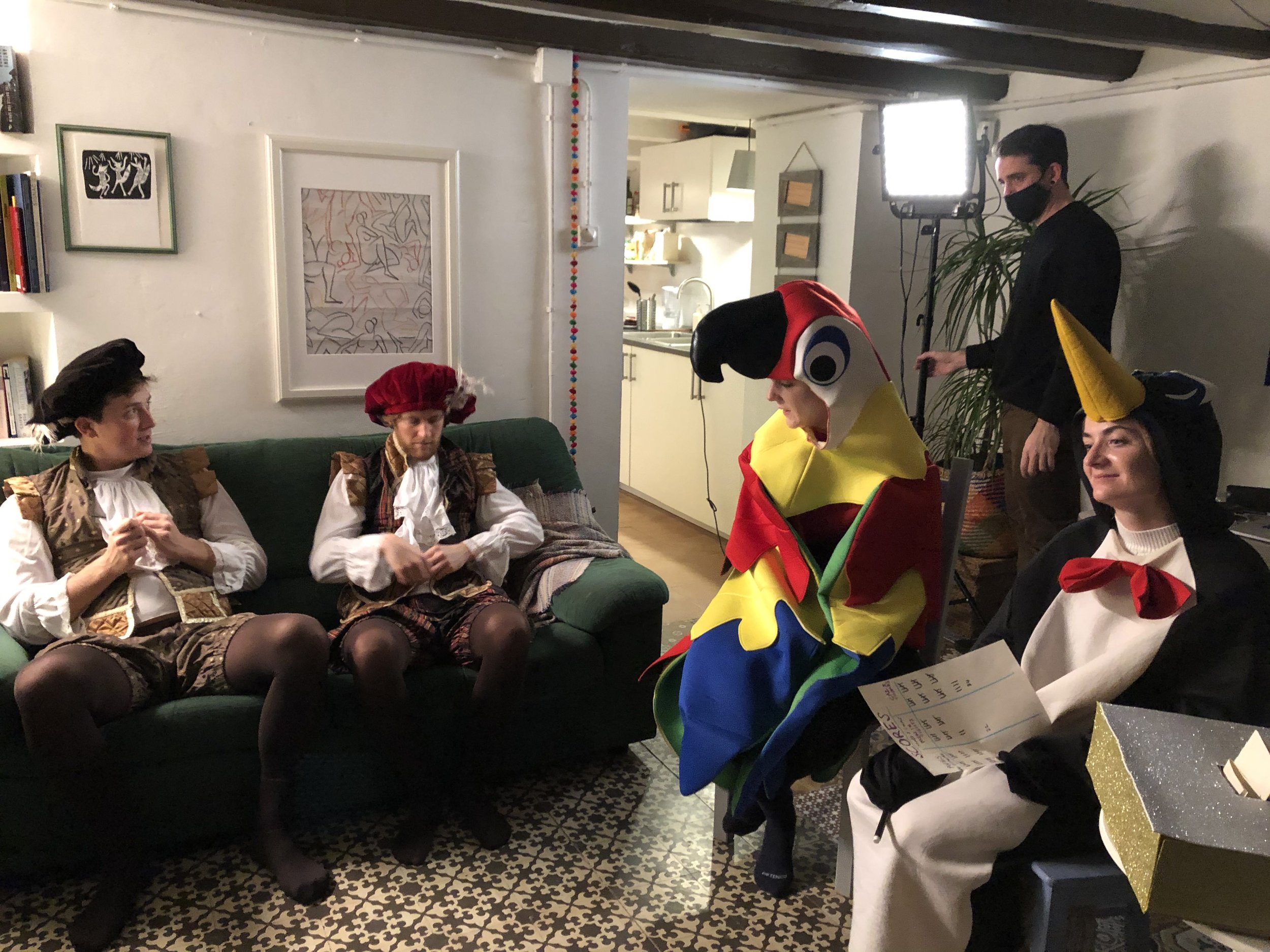 People in costumes gathered in a living room, appearing to be at a festive event or costume party. There are framed artworks on the wall, a bookshelf, and a person wearing a red, yellow, and blue clown or jester costume with a large hat, seated on a couch. Other individuals are dressed in historical or themed costumes, and there is someone standing near a bright studio light, possibly filming or photographing.