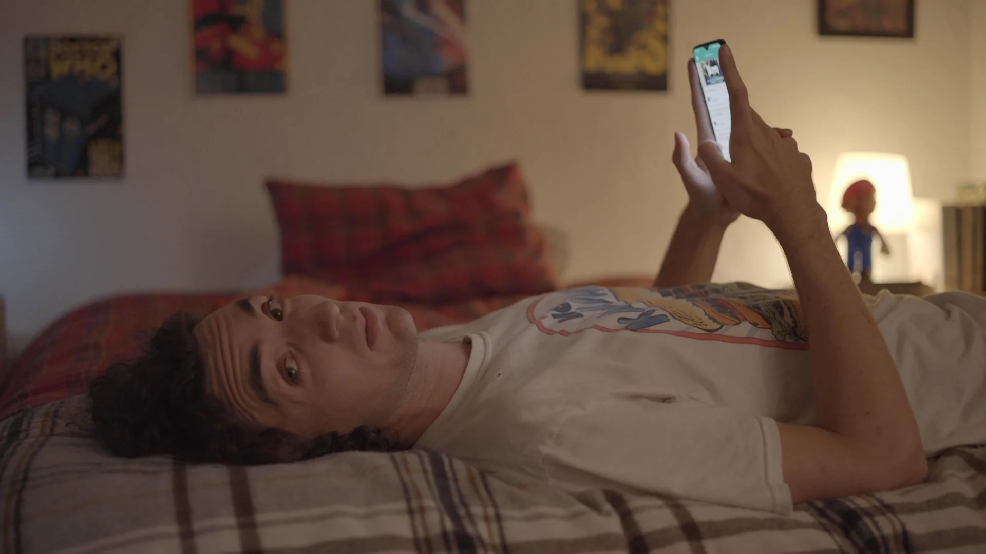 A young man lying on a bed, looking up at the camera, using a smartphone in a dimly lit bedroom with posters on the wall and a lamp on a bedside table.