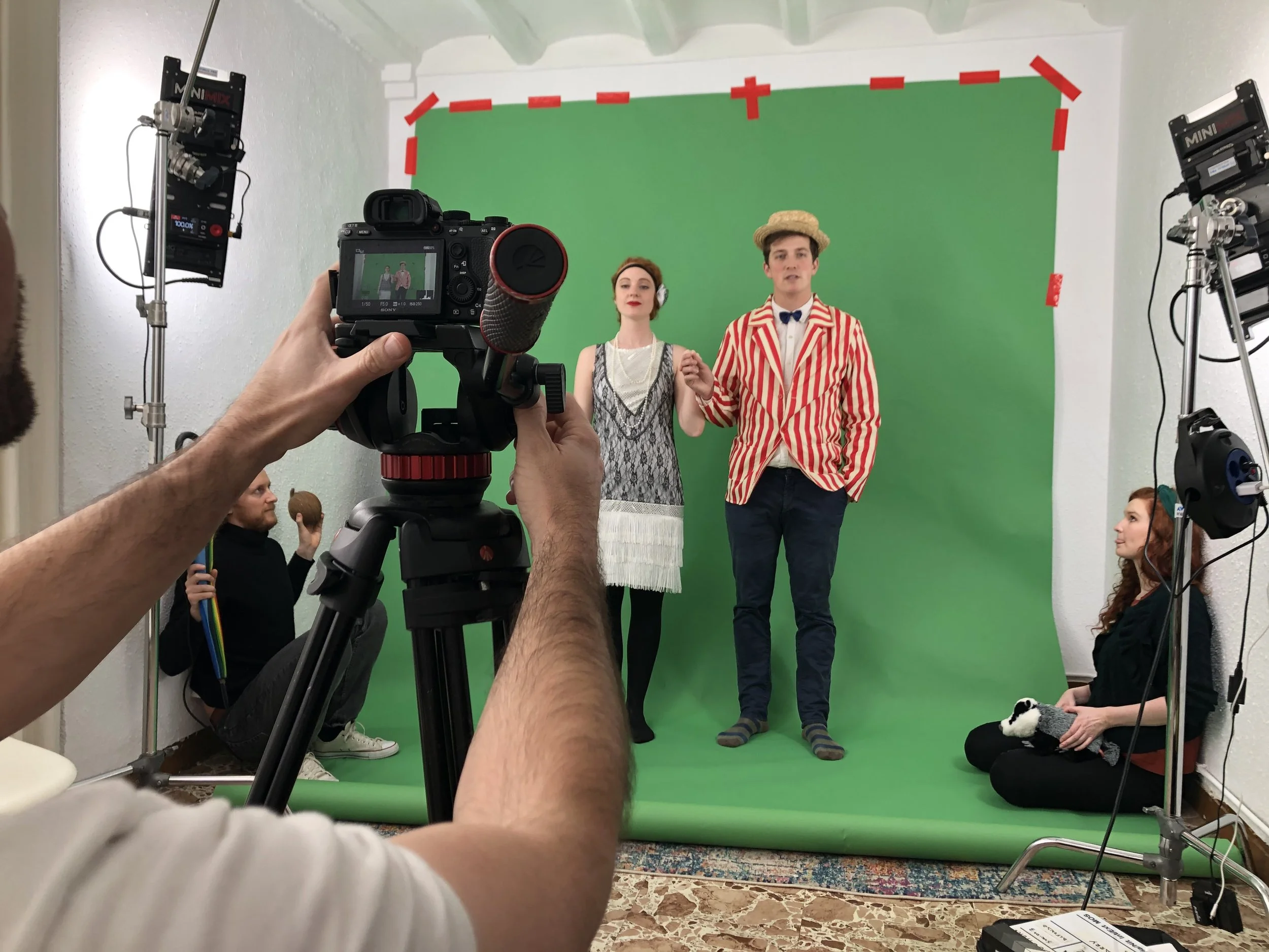 Actors in vintage costumes standing in front of a green screen during a photo shoot, with crew members and equipment around them.