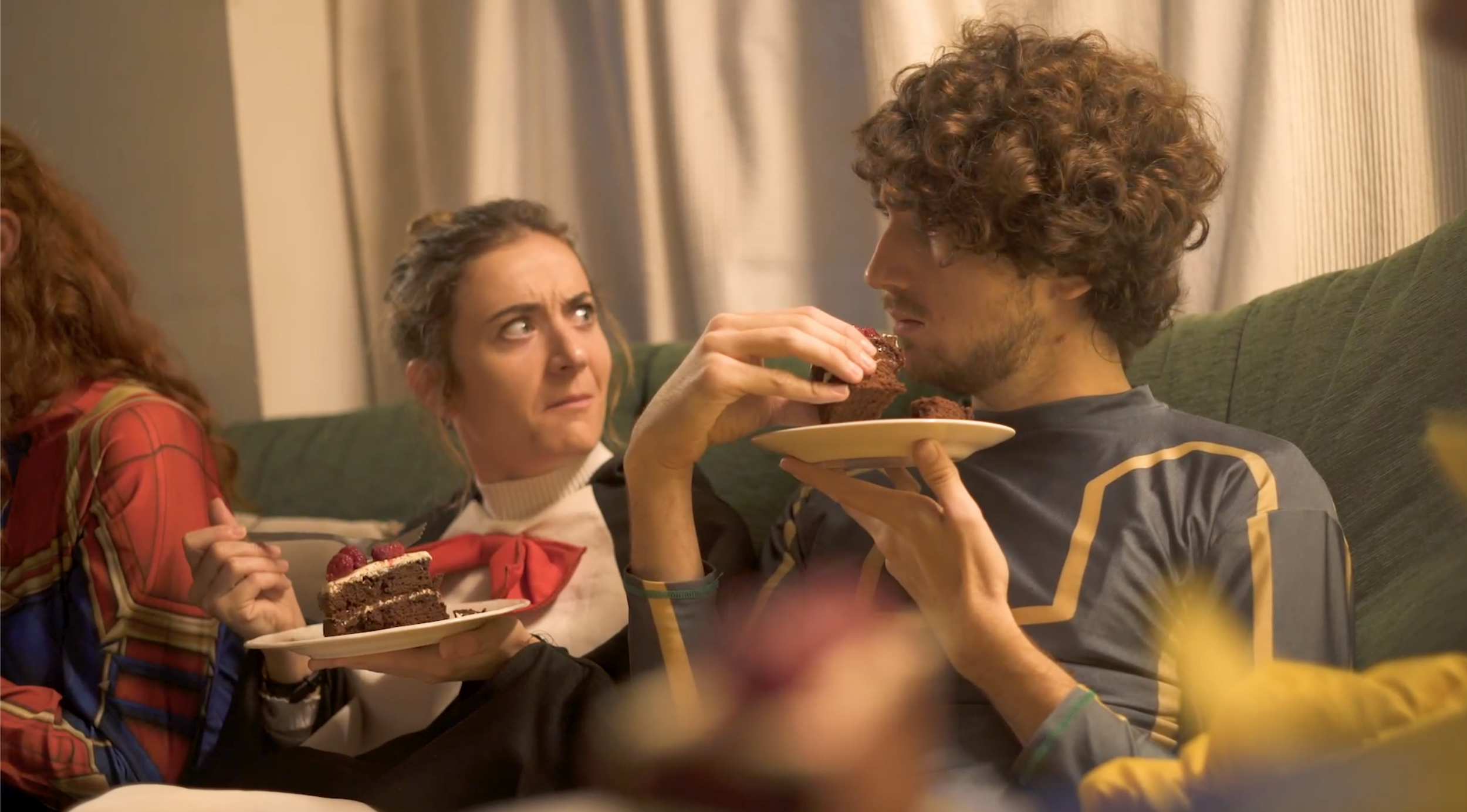 Three friends sitting on a couch, two women and one man, sharing a moment during a celebration with cake, in a cozy living room with curtains.