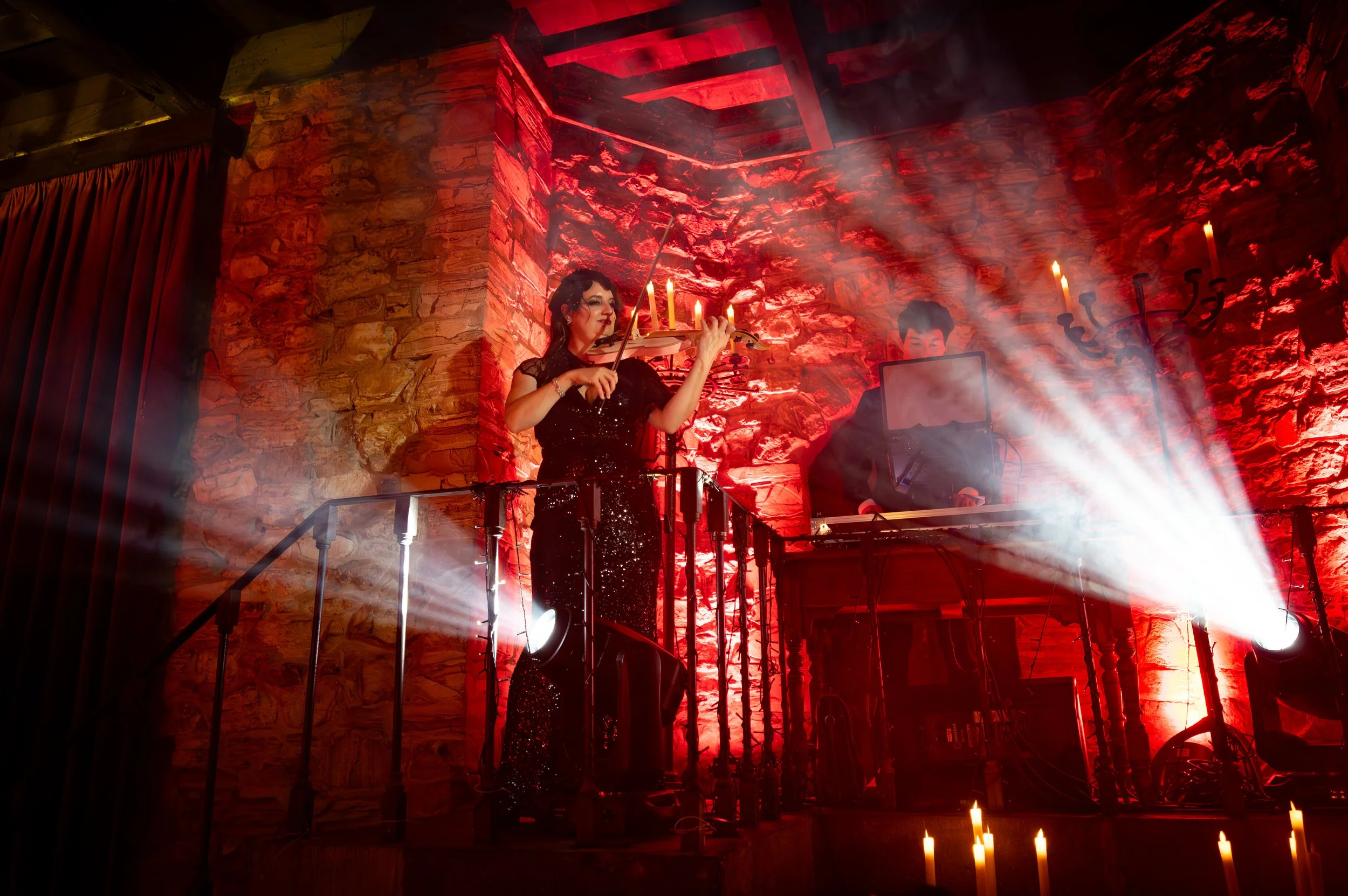A woman in a black sequined dress playing the violin on a stage illuminated with red lights and candles, with a person in the background operating electronic equipment.
