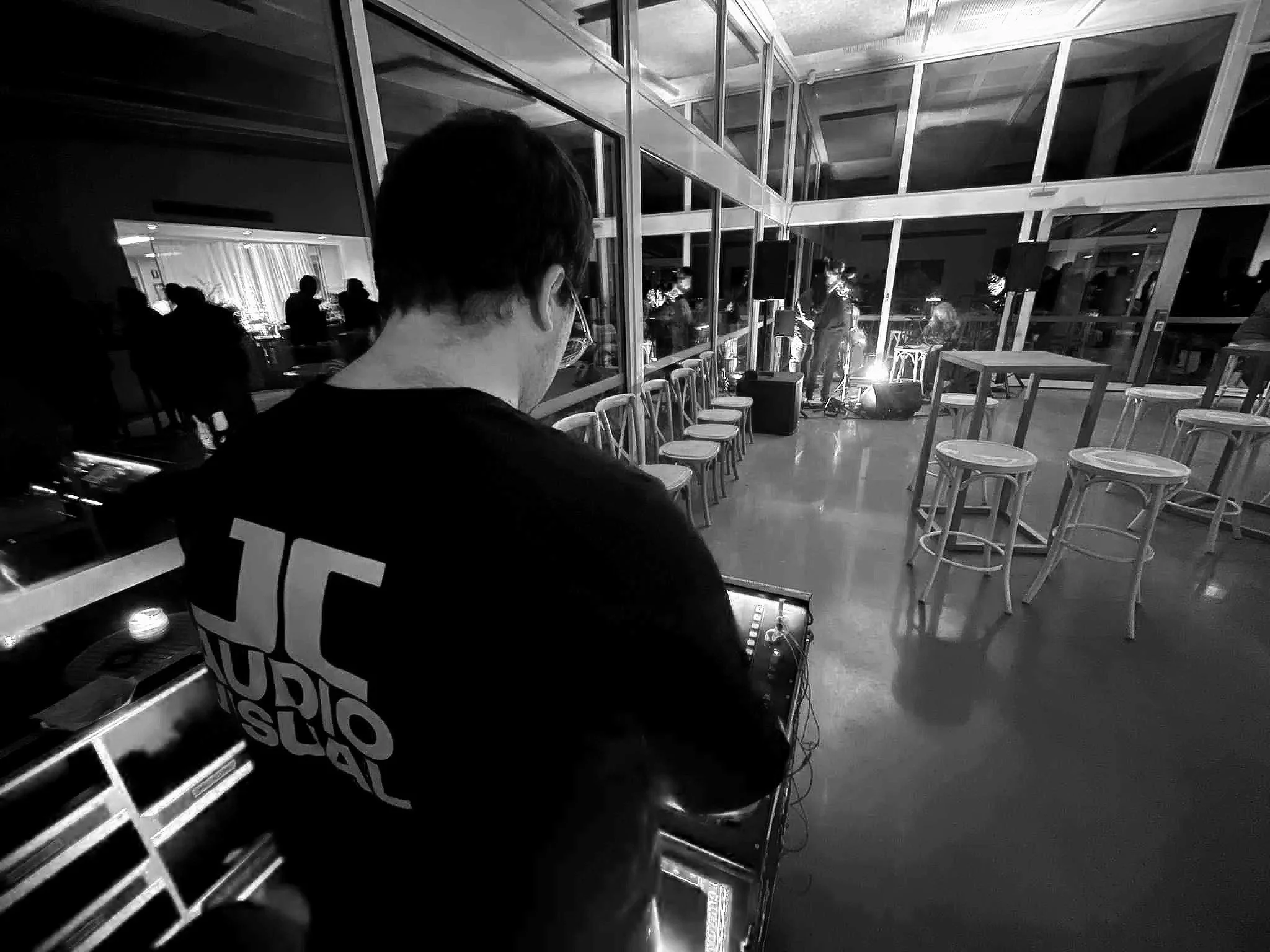 A person wearing a black shirt with 'JC Studio' printed on the back is operating DJ equipment in a large indoor space with glass walls. Several people are gathered in the background, some appear to be performing on a small stage with bright lighting. Empty chairs are lined up along the glass wall, and the room has a modern design with high ceilings and reflective floors.
