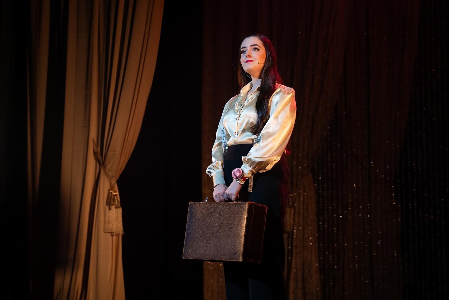 A woman with dark hair and light skin stands on stage, holding a briefcase in front of her. She wears a shiny silk blouse, dark pants, and has a headset microphone. The stage has golden curtains on the left and a dark, sparkly backdrop.