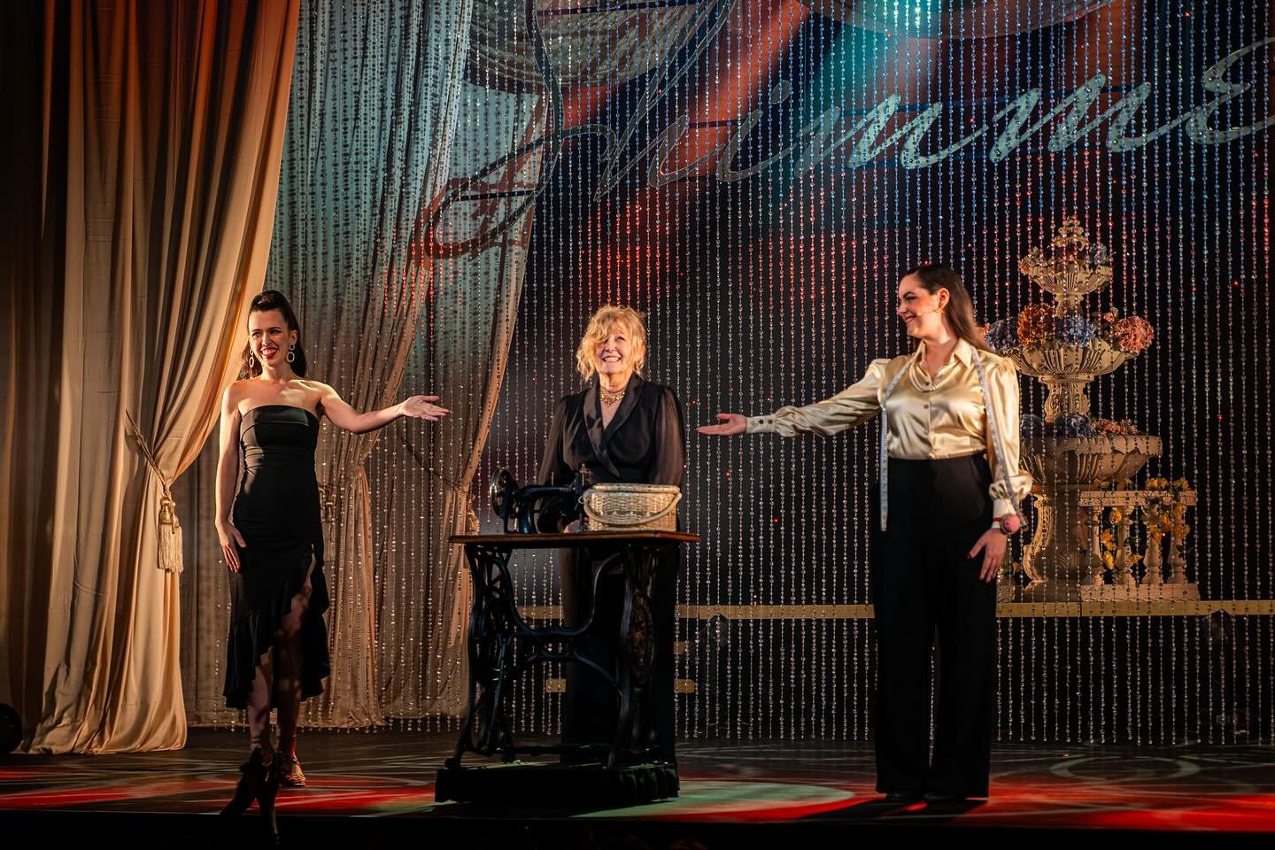 Three women on stage with golden curtains and beaded backdrop. One woman in a black dress, one in a shiny blouse, and an older woman with blonde hair in a black outfit. They are smiling and gesturing towards each other during a performance or presentation.