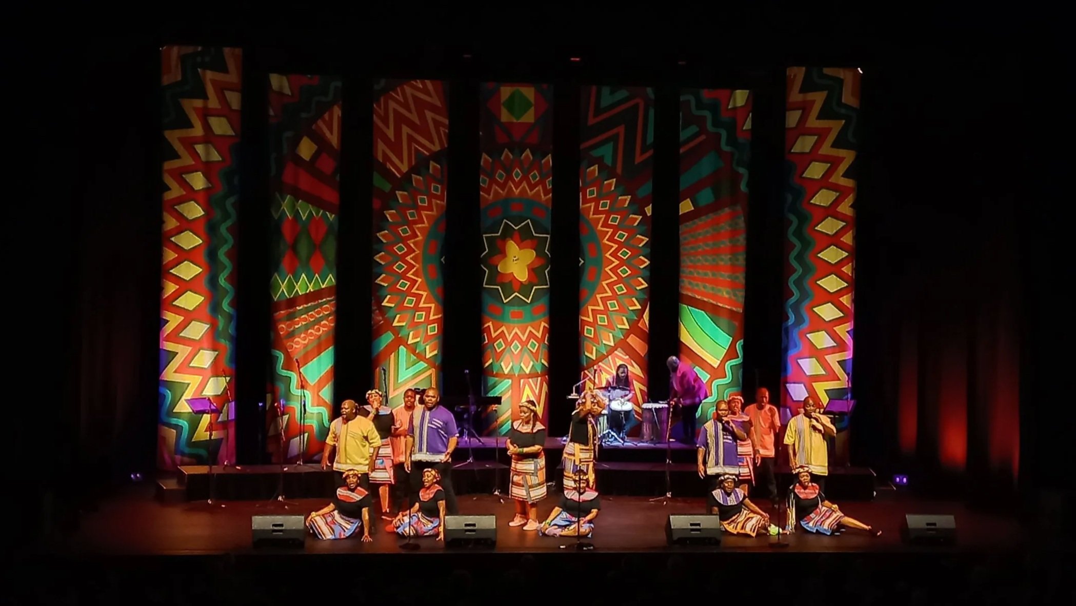 A stage performance featuring a group of performers wearing colorful traditional African attire. Behind them are vibrant, geometric patterned backdrops. Some performers are seated on the floor, while others stand with microphones, and a musician is playing drums in the background.