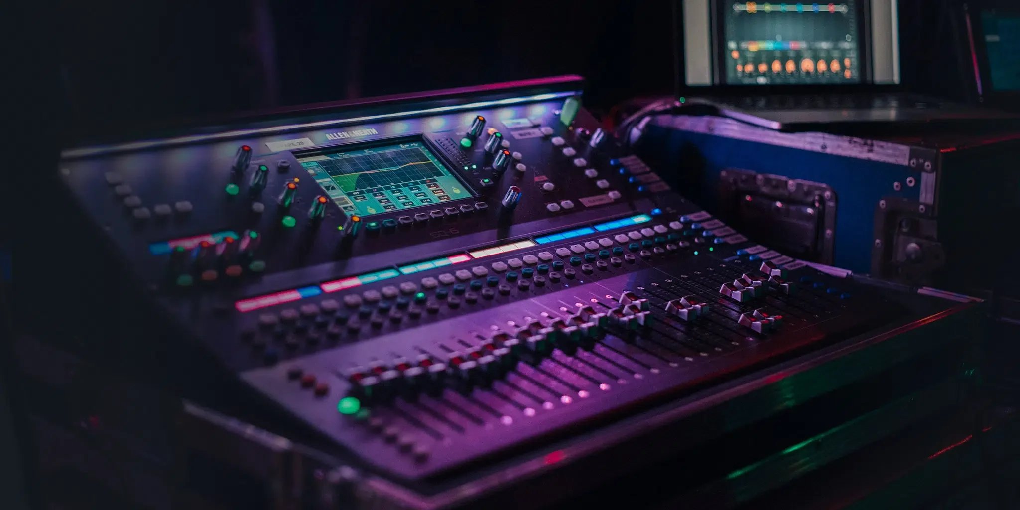 A professional audio mixing console with colorful lights, faders, and knobs, illuminated in a dark setting, with a laptop displaying audio editing software nearby.