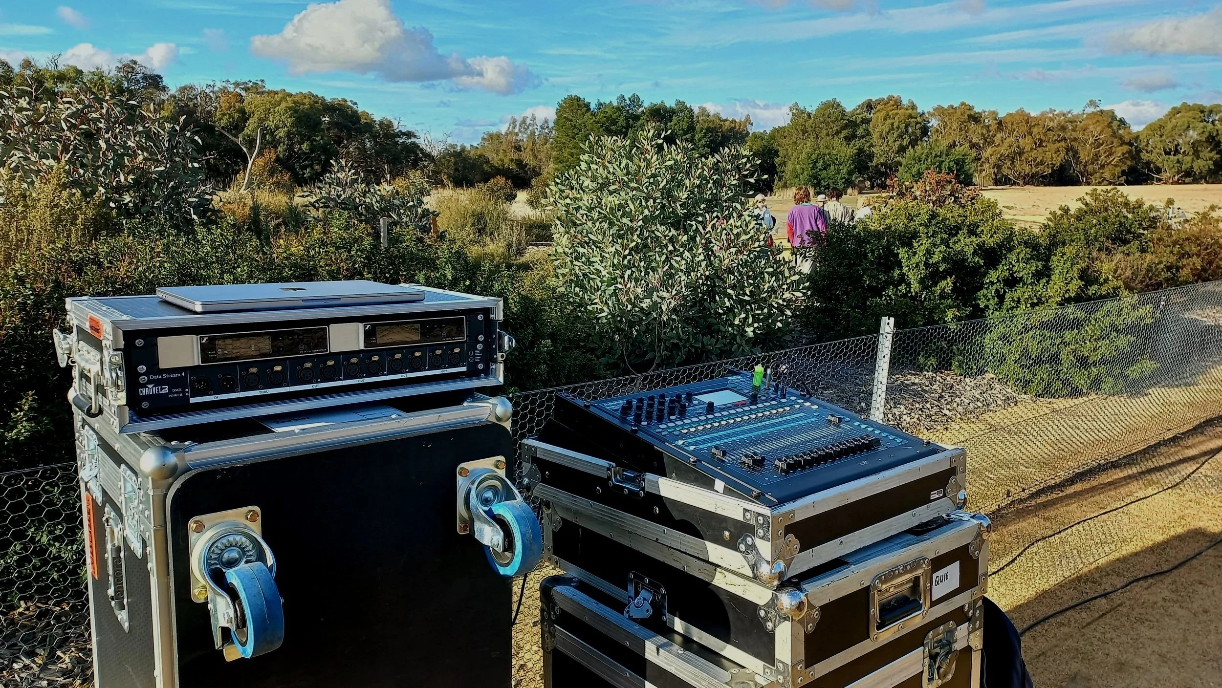 Outdoor scene with DJ equipment on tables, including a DJ mixer and a speaker case, set against a backdrop of trees and bushes under a partly cloudy sky, with a few people walking in the distance.