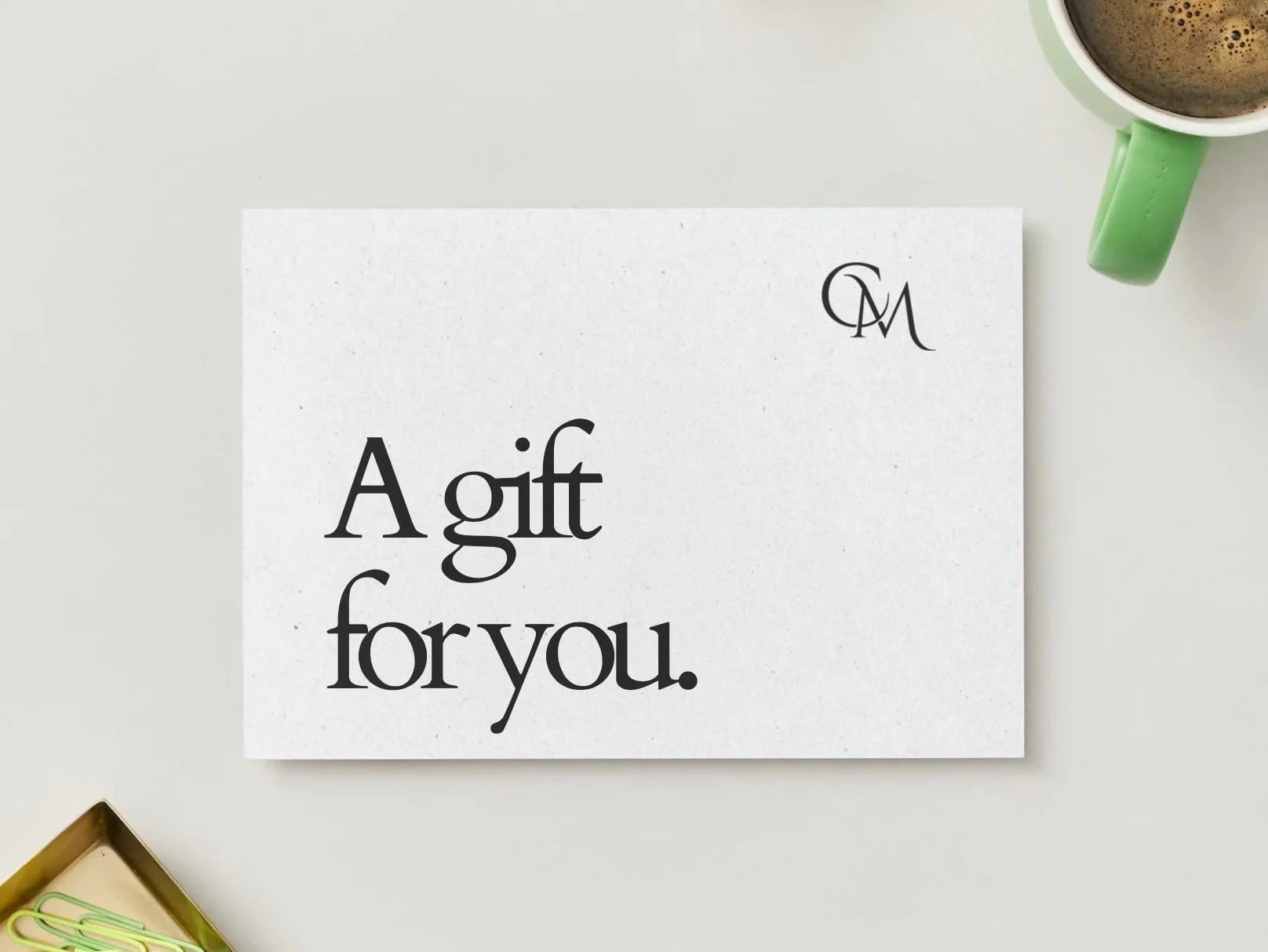A white greeting card with the message "A gift for you." in black text, placed on a white surface, with a green mug of coffee in the top right corner and a small partially visible tray with paper clips in the bottom left corner.