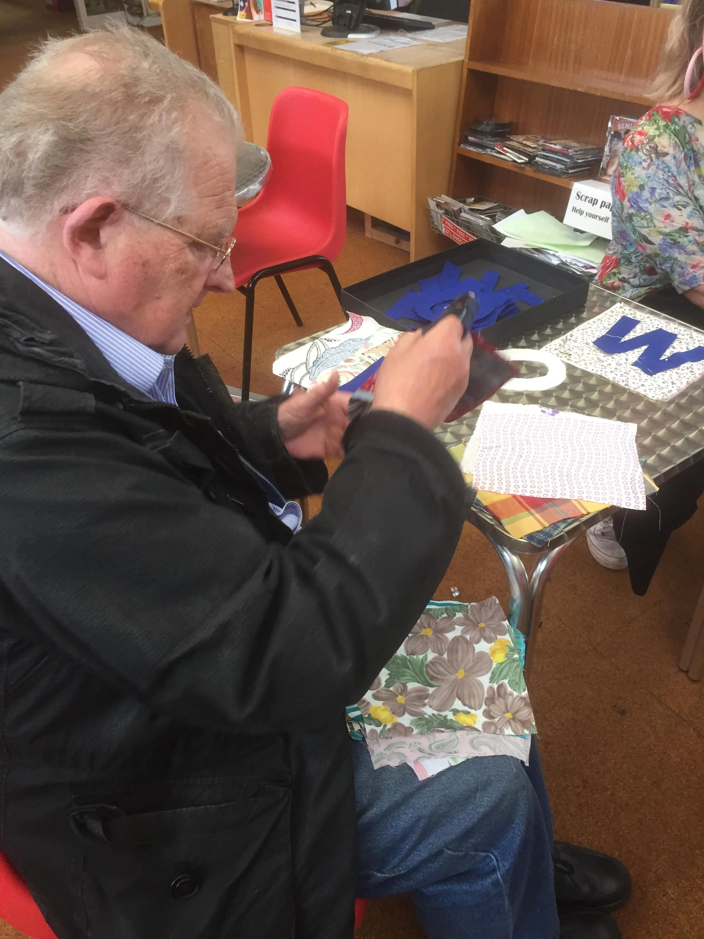 An elderly man with glasses and gray hair is sitting at a table, sewing fabric with floral patterns. The table has various fabric pieces and crafts materials, with a background of a wooden bookshelf and a sign that reads 'Scrap paper.'