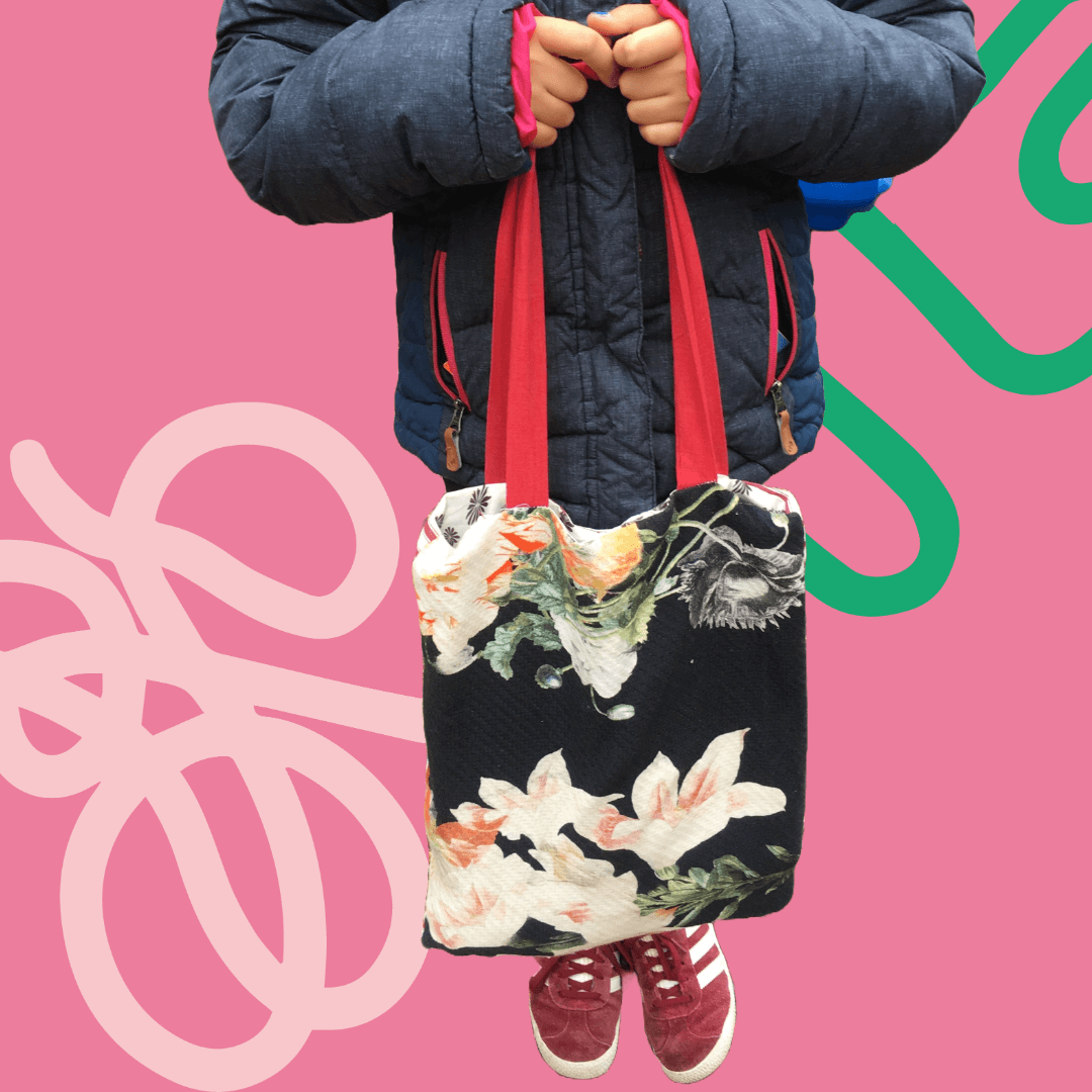 Person holding floral tote bag with red straps, wearing a blue jacket and pink shoes on a pink background.