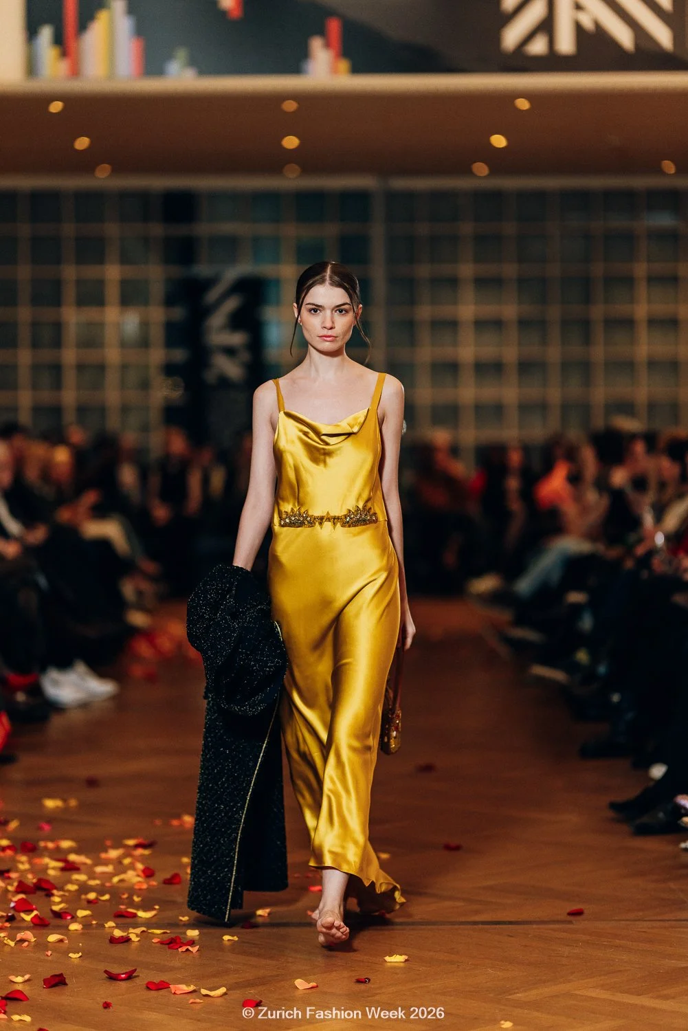 Ein Model läuft auf dem Laufsteg während der Zurich Fashion Week 2025. Sie trägt ein gelbes Kleid mit schwarzen Streifen, darüber eine gelbe Trage und ein Kabel um das Bein.