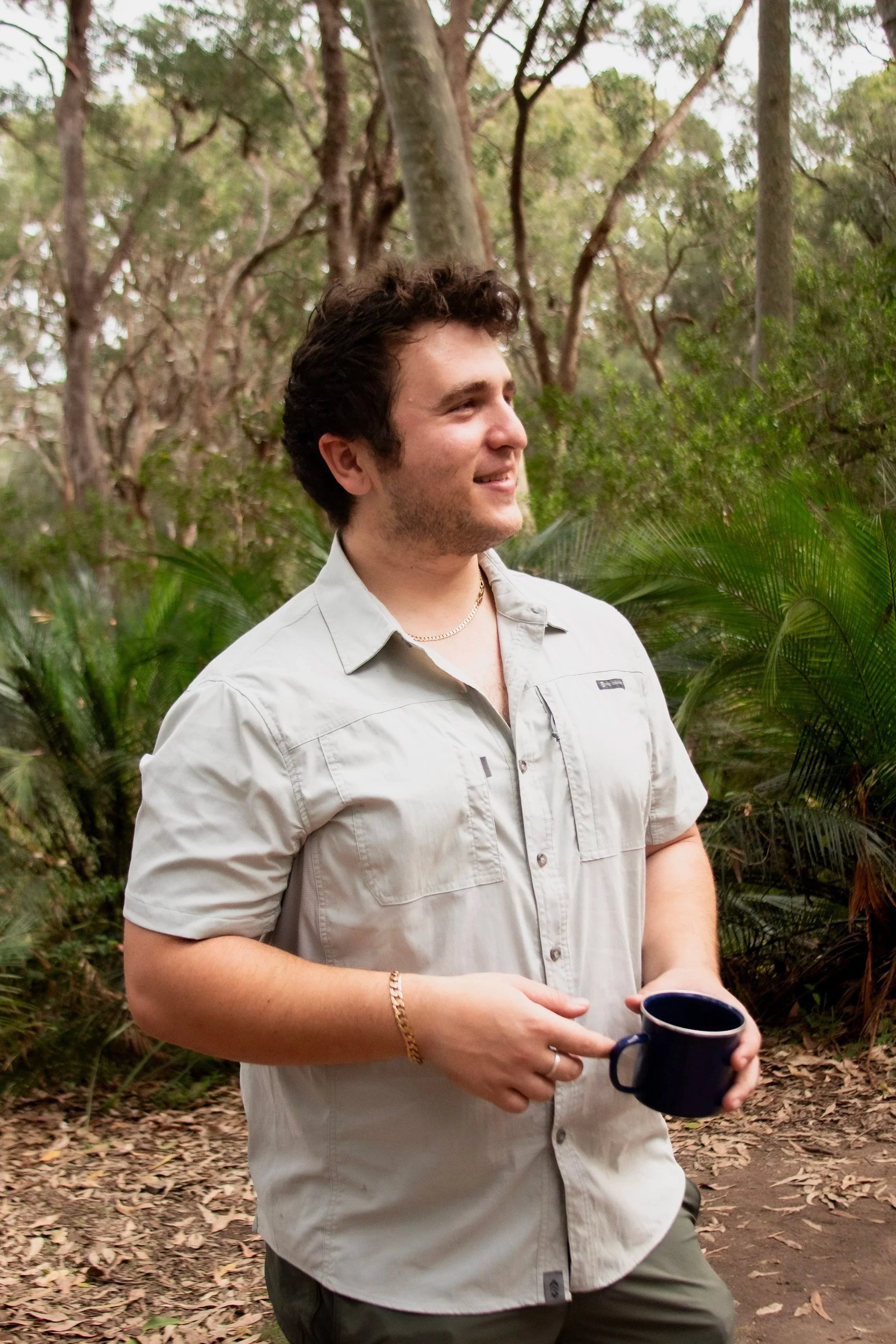 A young man with dark, curly hair wearing a beige short-sleeve button-up shirt and holding a blue mug, standing outdoors in a wooded area with trees and leaves in the background.