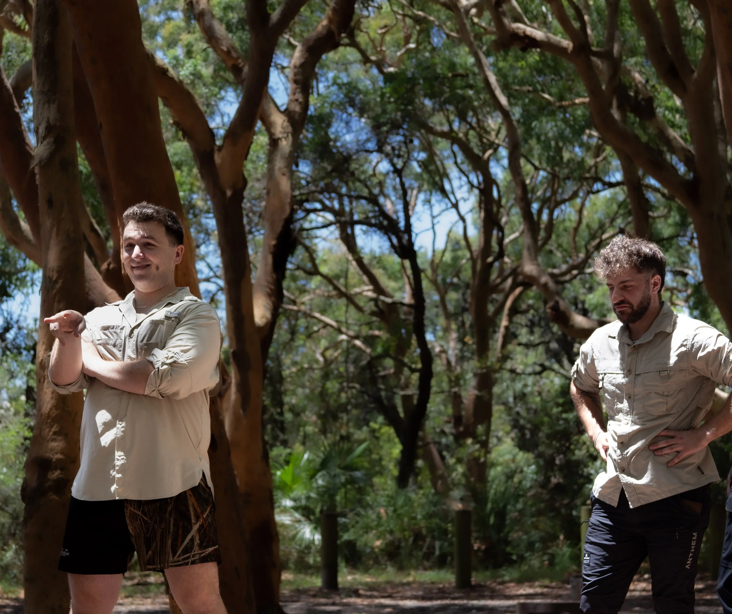 Two men standing outdoors among tall trees with green leaves, facing slightly to the side. One man on the left has short dark hair and is smiling, wearing a light-colored shirt with rolled-up sleeves. The man on the right has curly hair and a beard, looking downward, wearing a light-colored shirt.