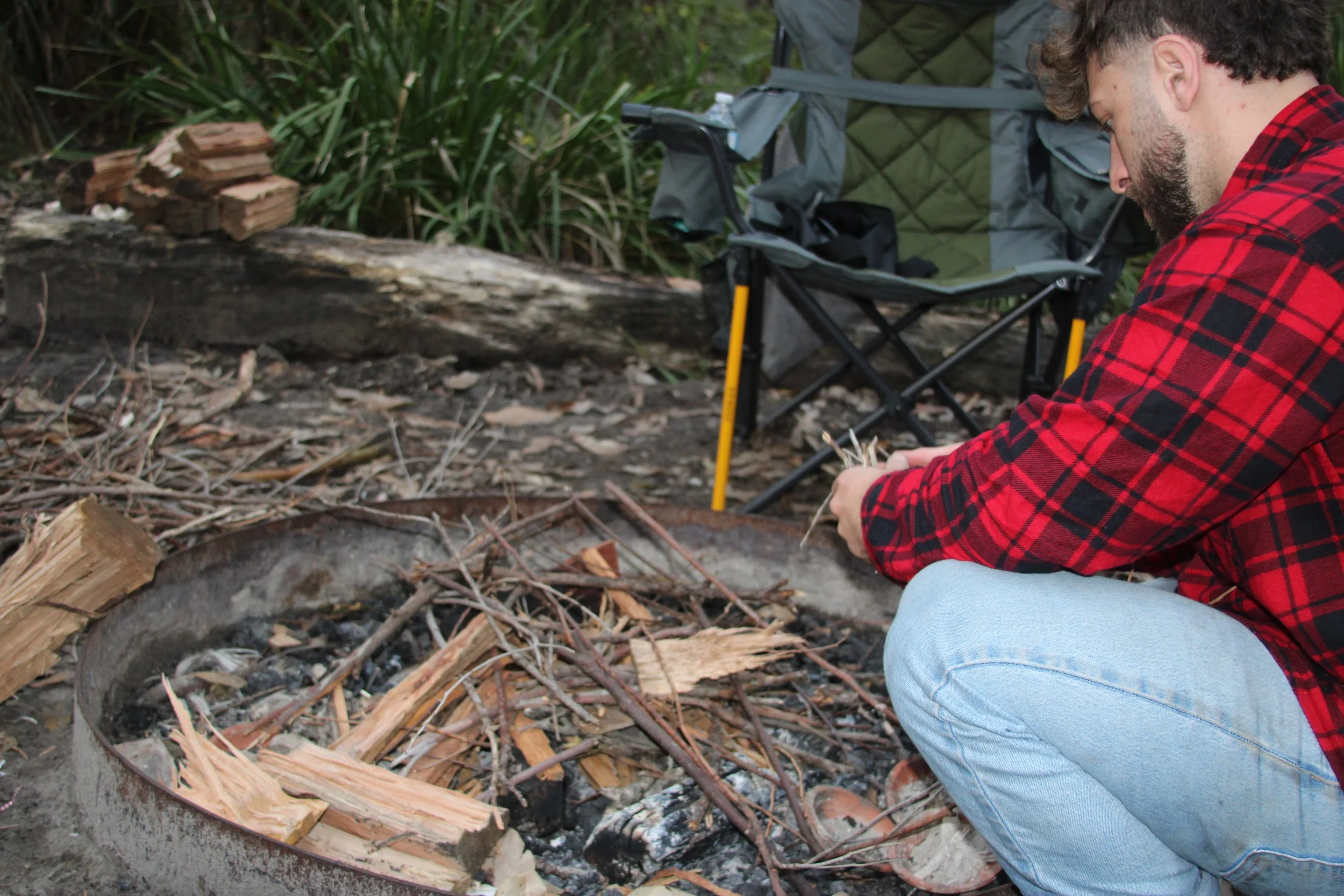 A man in a red plaid shirt crouches next to a campfire ring, preparing kindling and tinder for a fire in an outdoor setting. A camping chair is in the background.