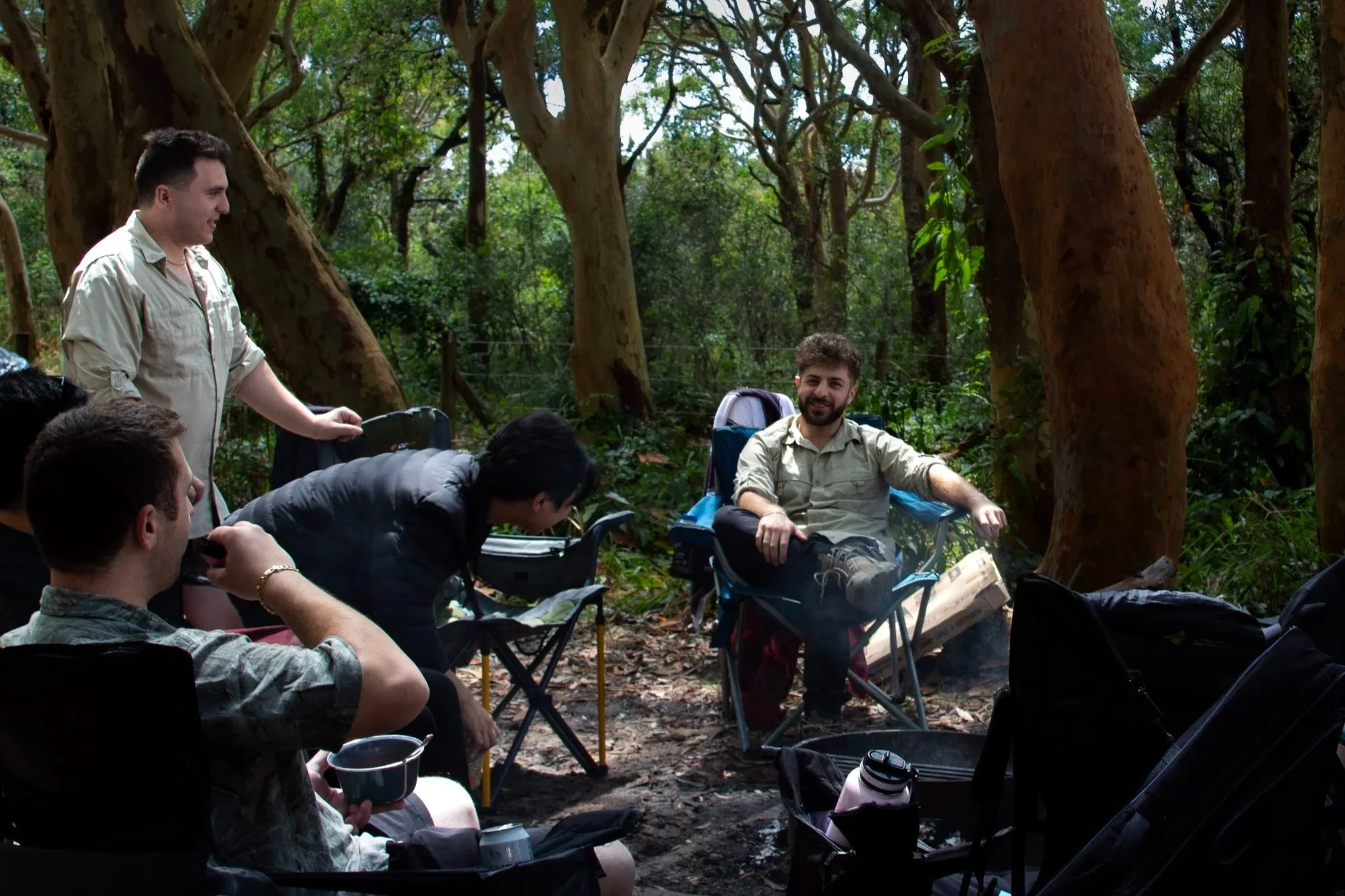 Group of people enjoying a camping trip in a forest, sitting around a fire with trees surrounding them.