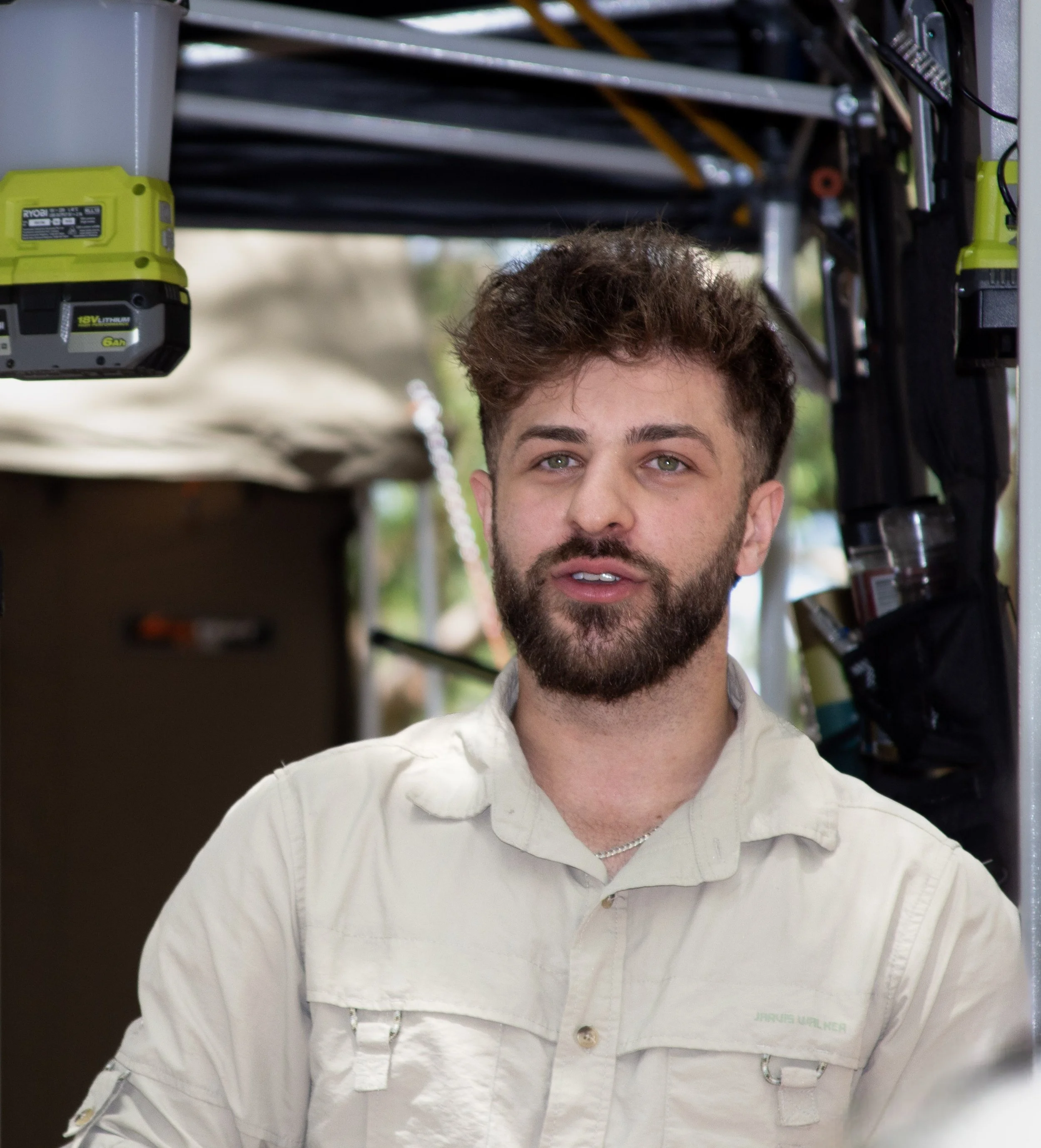 A young man with a beard and light brown hair is outdoors, wearing a light-colored shirt, standing in front of a metal structure and some tools, possibly in a workshop or outdoor workspace.