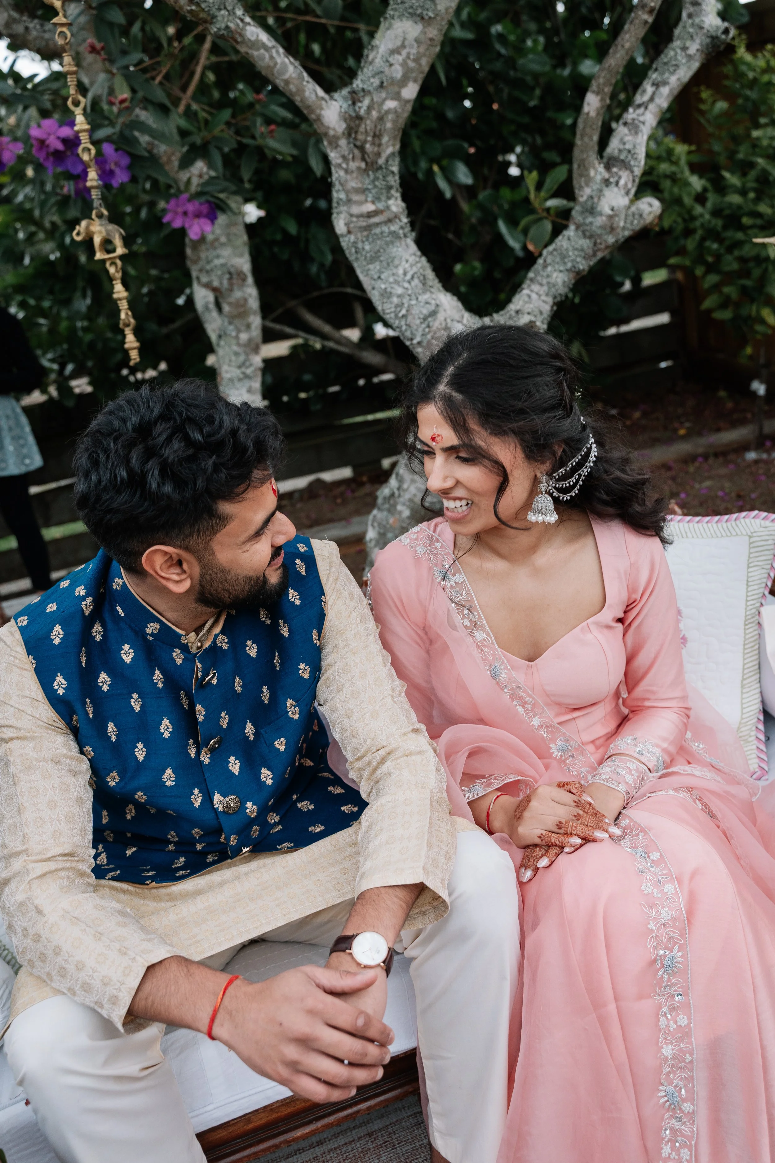 A man and a woman dressed in traditional Indian attire sitting outdoors, smiling and looking at each other.