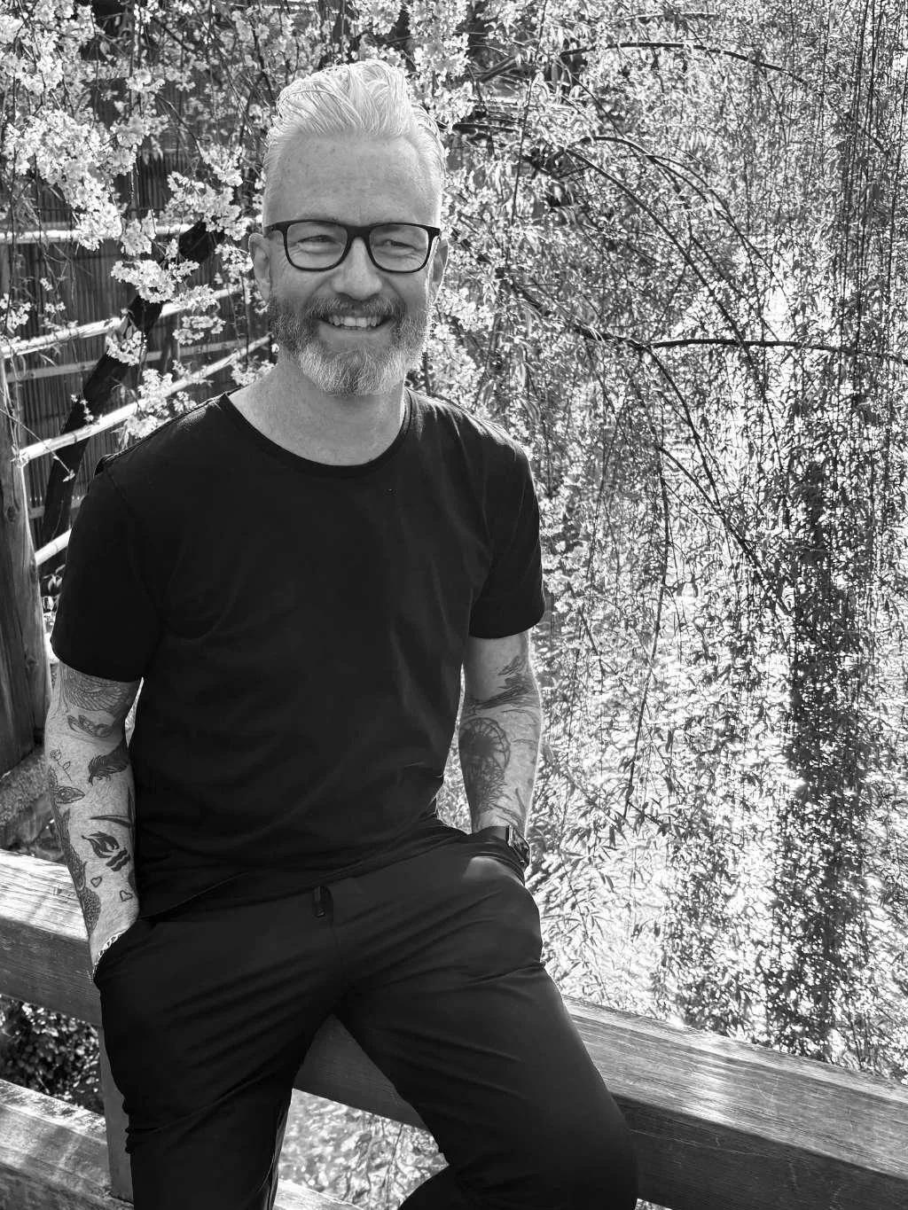 A smiling man with glasses and a beard sitting outdoors on a bench, with trees and flowering bushes in the background.