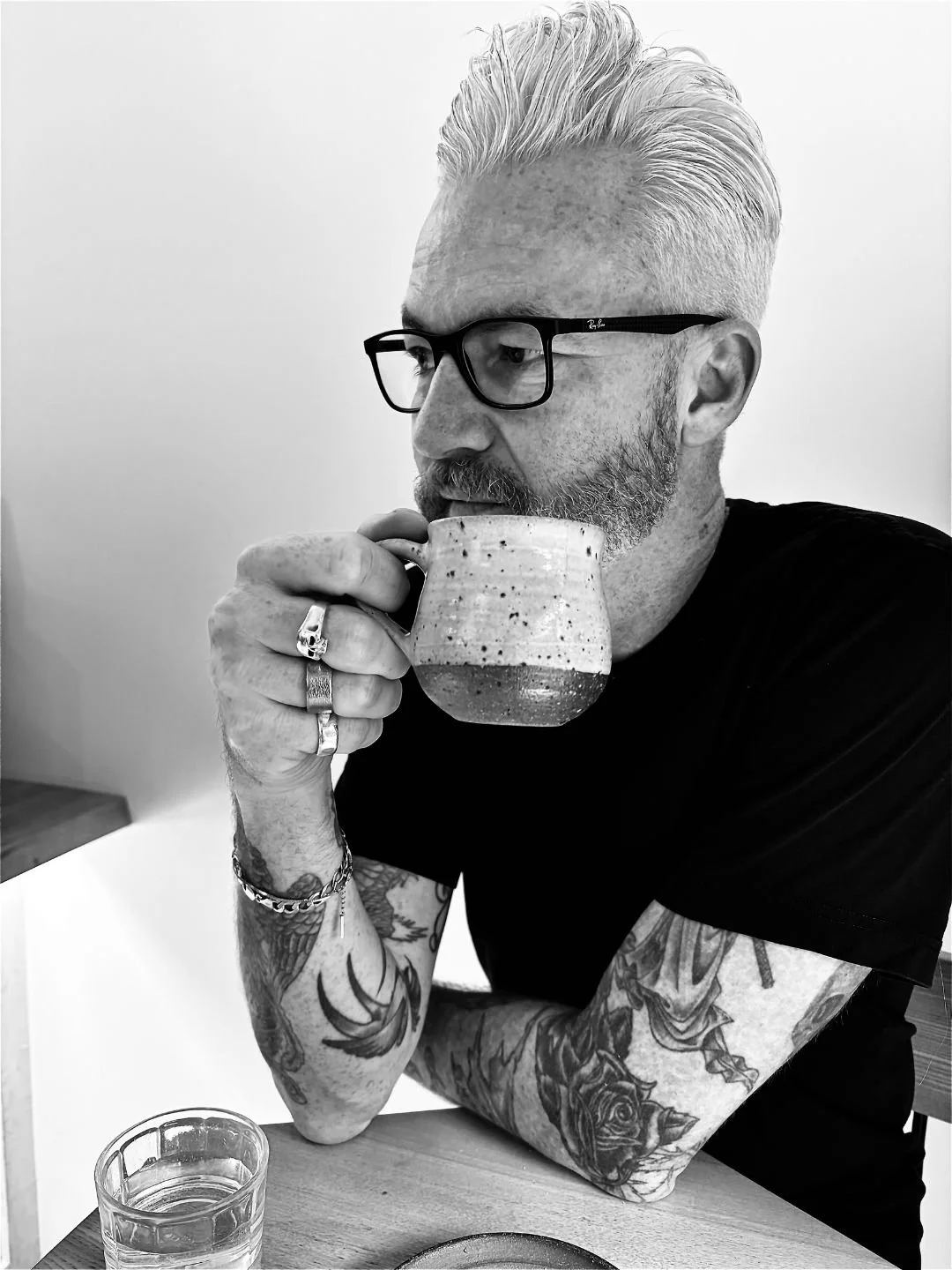 A man with tattoos and glasses sipping from a ceramic mug at a table with a glass of water.