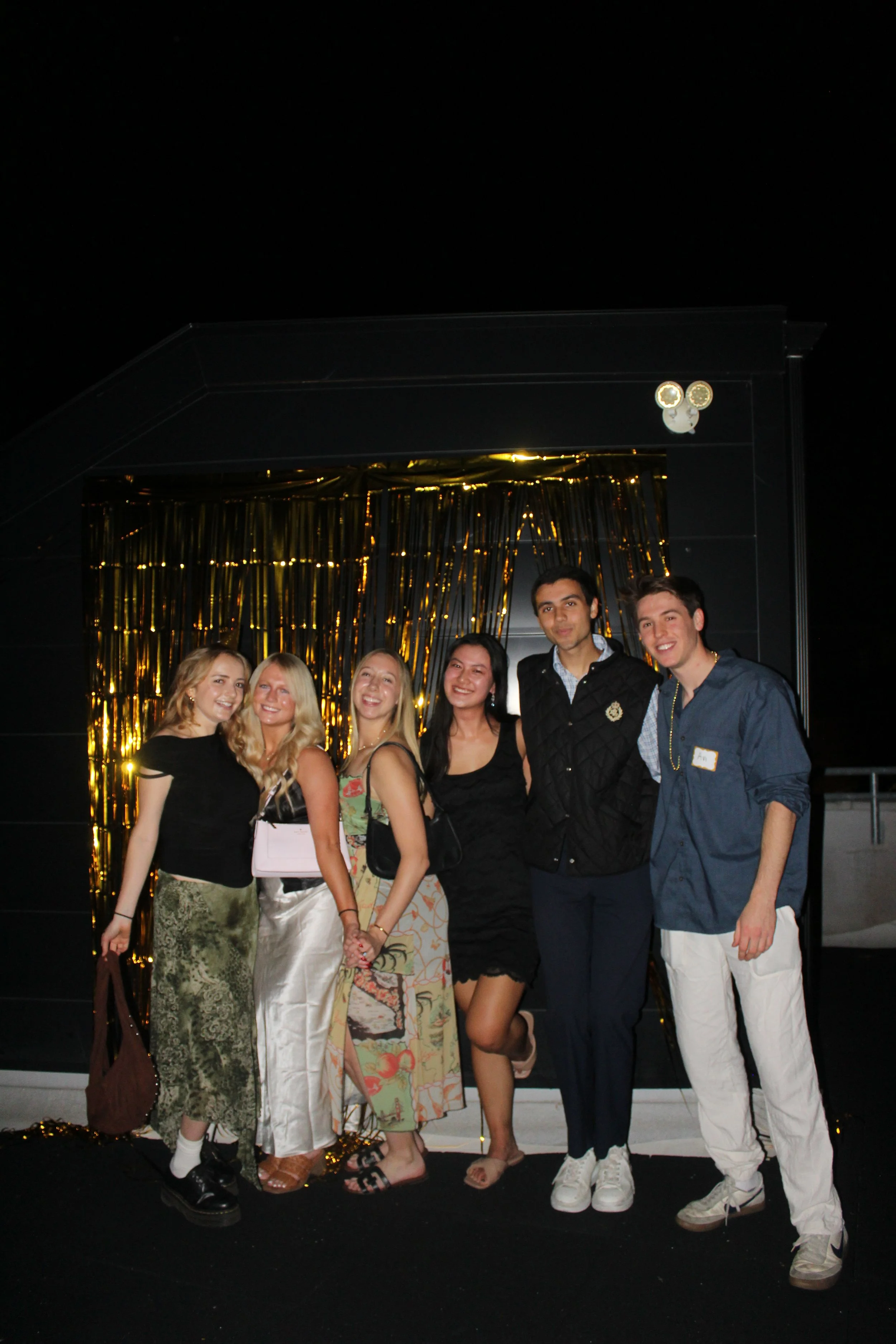 Group of six young adults posing in front of a black backdrop with gold curtains, at night.