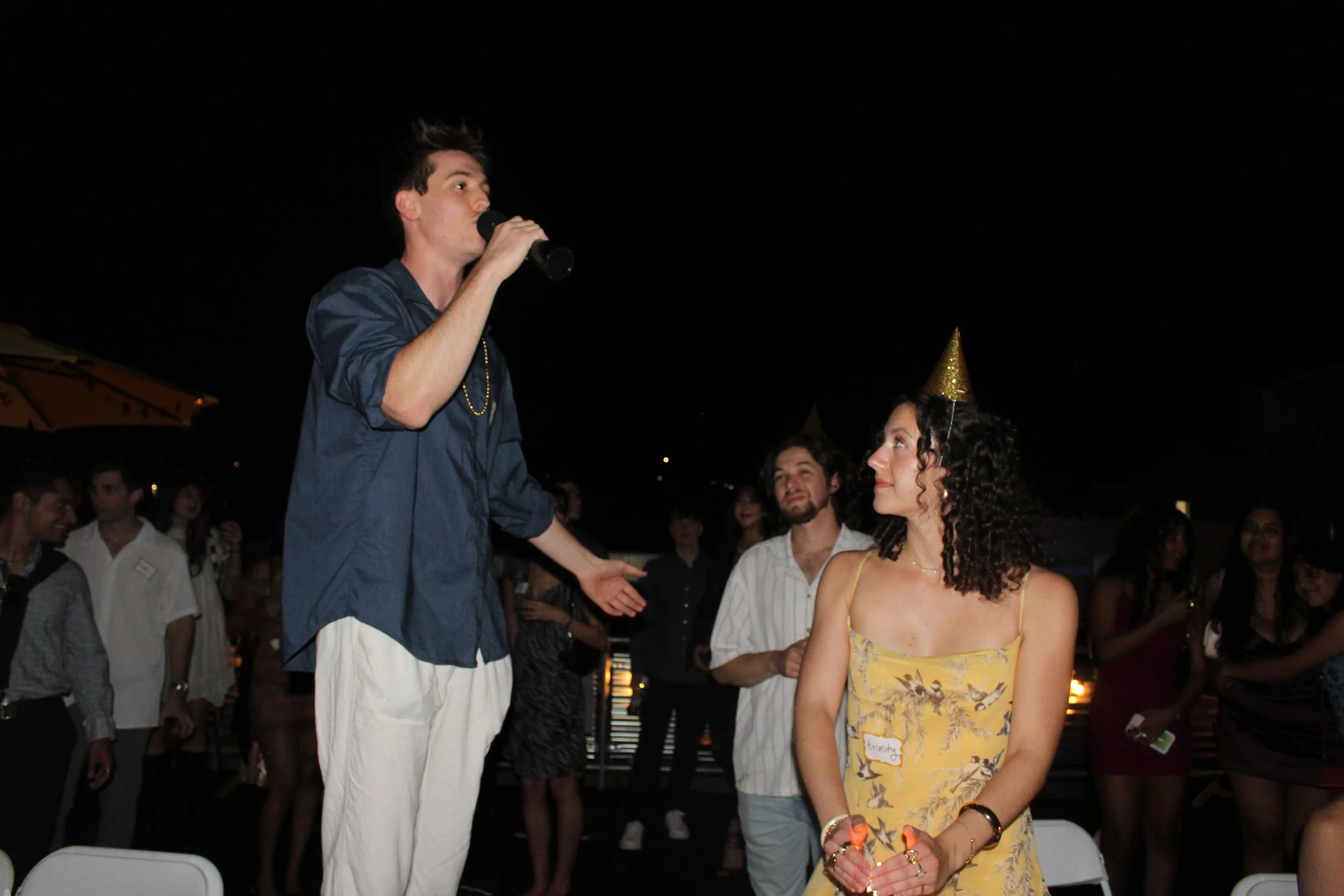A man speaking into a microphone at a nighttime celebration with a woman wearing a gold party hat, decorated with a tropical-print dress, and surrounded by other people at an outdoor party.