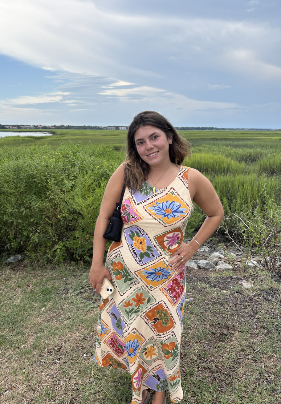 Young woman wearing a colorful maxi dress with floral and country-themed patches, standing outdoors near green marshland with a cloudy sky in the background, holding a phone and smiling.