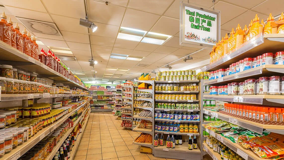 A well-stocked aisle in an Asian grocery store with various sauces, condiments, and packaged food products on shelves. A sign overhead in multiple languages indicates Thai products.