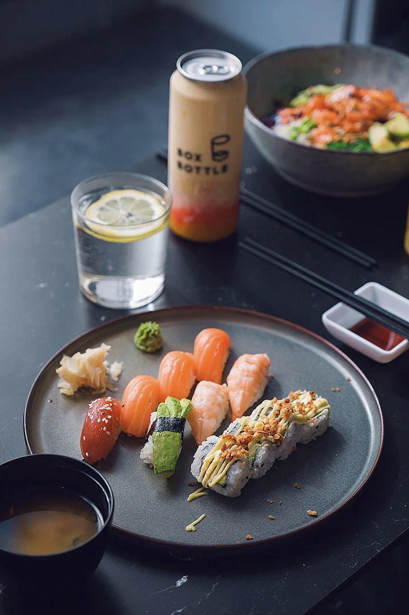 Plate of assorted sushi with salmon, shrimp, and avocado, accompanied by wasabi and ginger, next to a canned beverage and a bowl of salad.