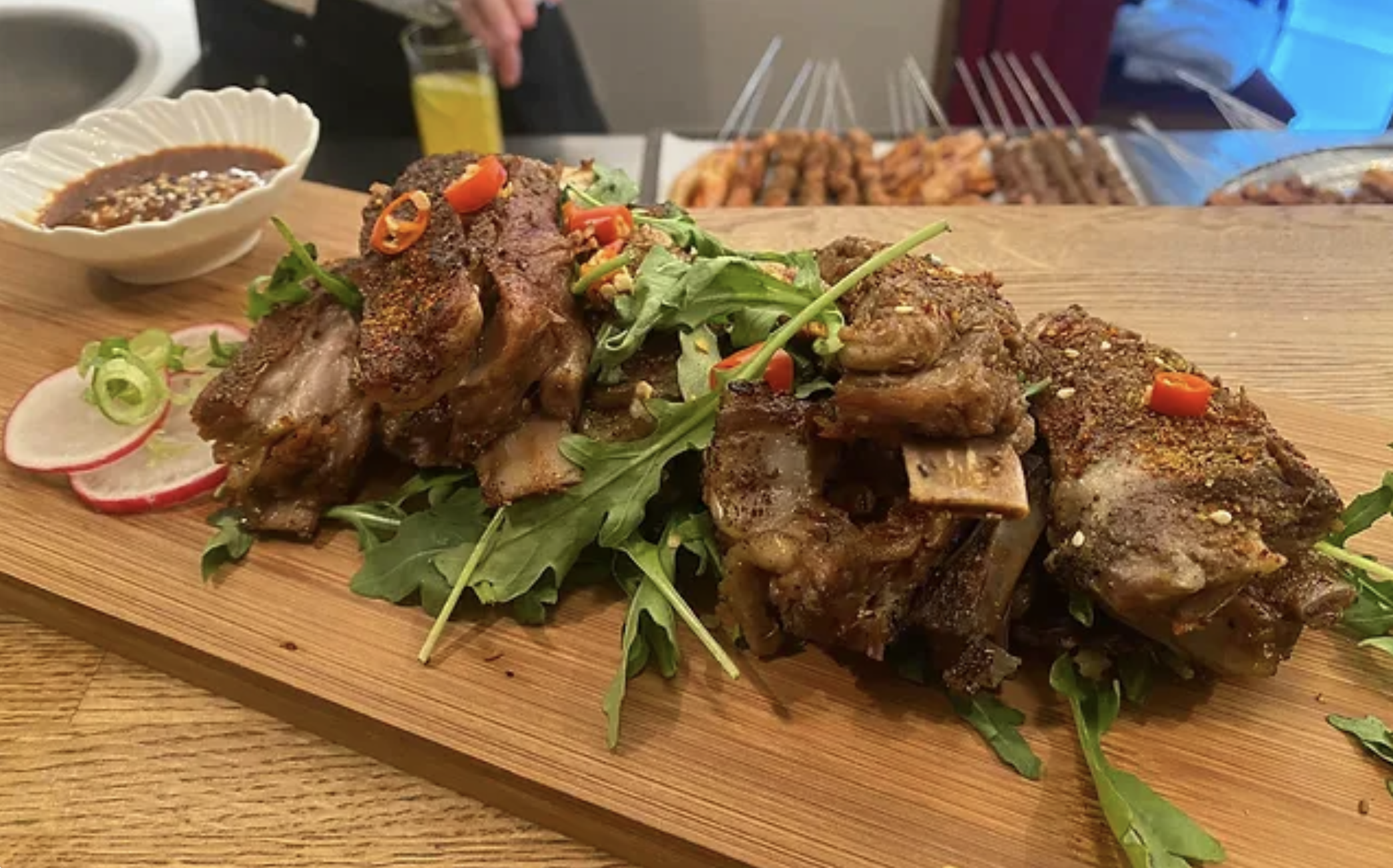 Grilled meat ribs with arugula and sliced radishes on a wooden board, accompanied by a bowl of dipping sauce.