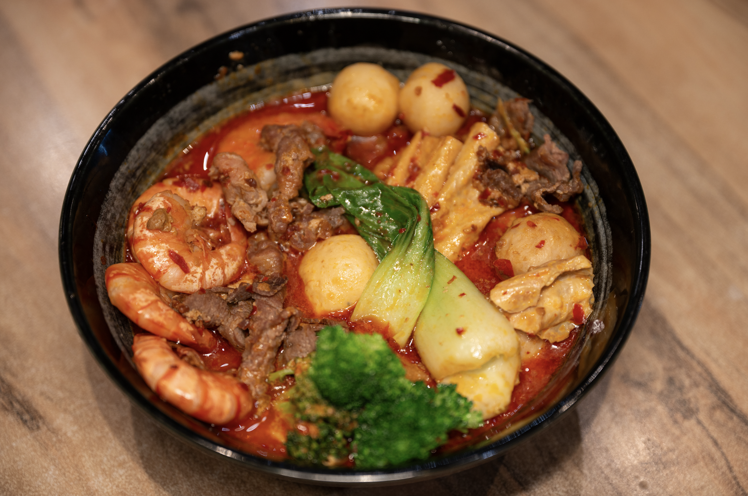 A bowl of spicy Asian-style soup with shrimp, beef, bok choy, tofu, broccoli, and quail eggs in a red broth.