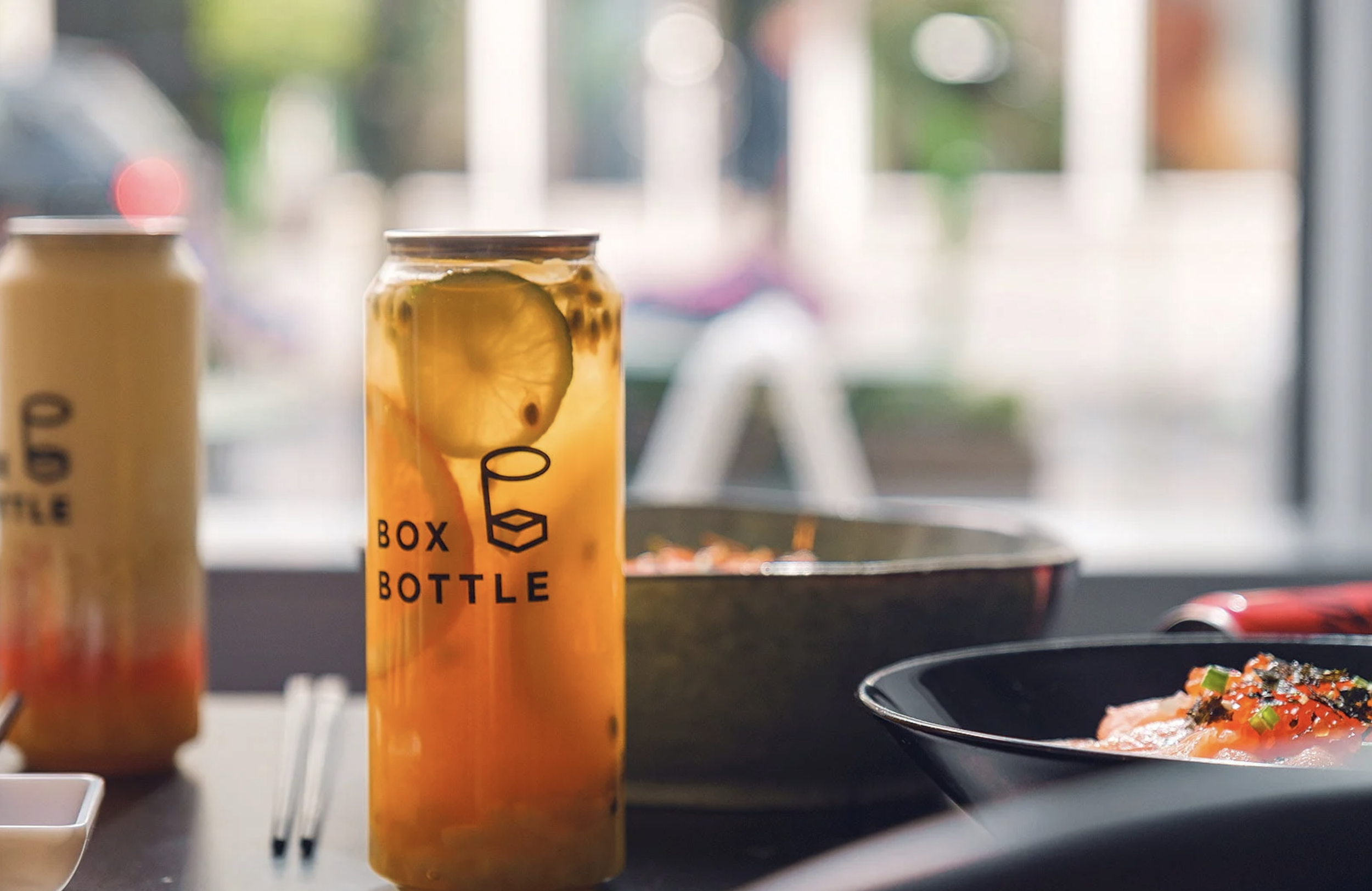 Close-up of a passion fruit lemonade in a clear plastic bottle labeled 'Box Bottle,' with lemon slices and passion fruit seeds, next to bowls of food, on a table near a window.