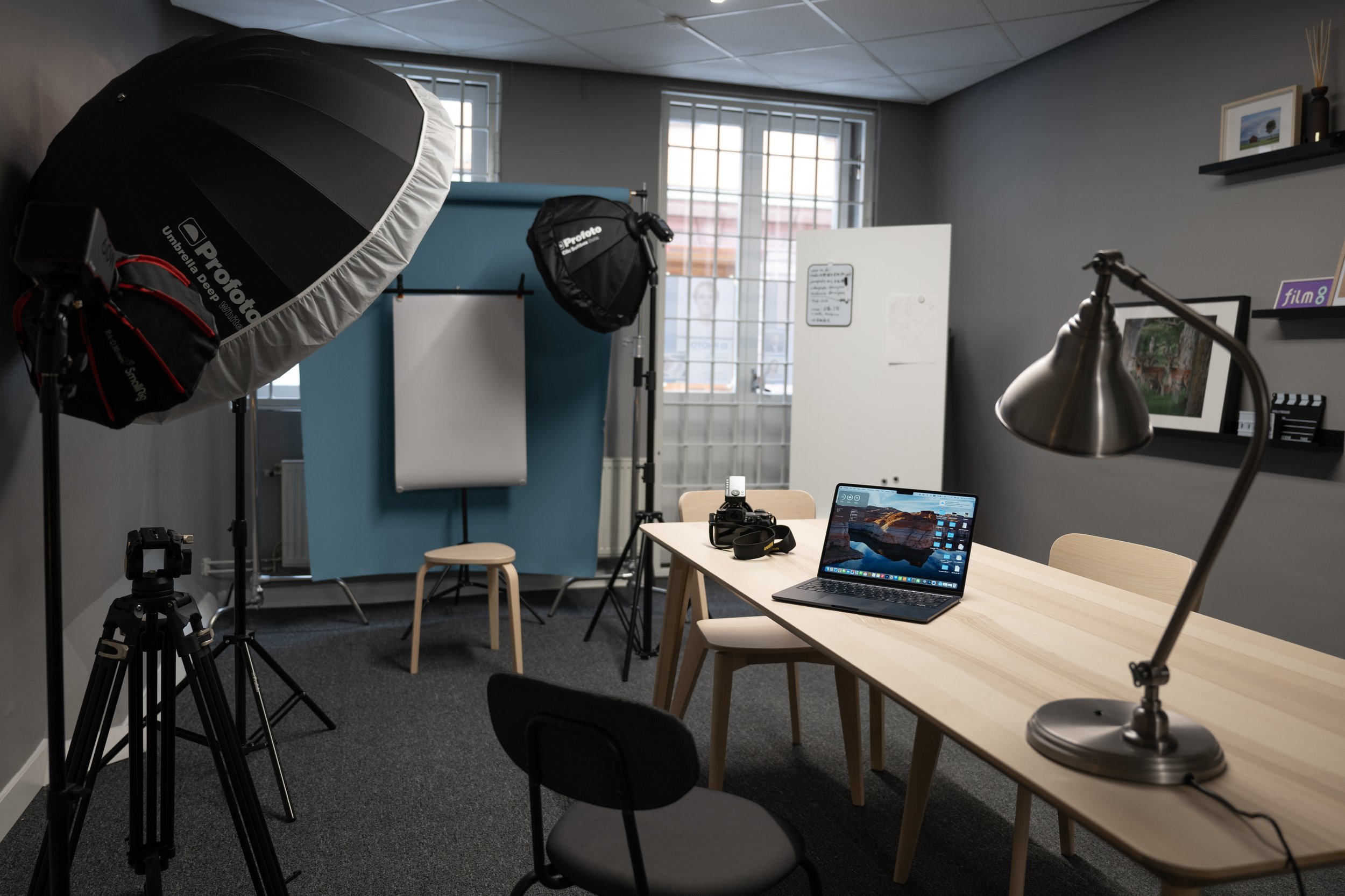 Photography studio setup with lighting equipment, backdrop, camera on tripod, laptop, and office desk.