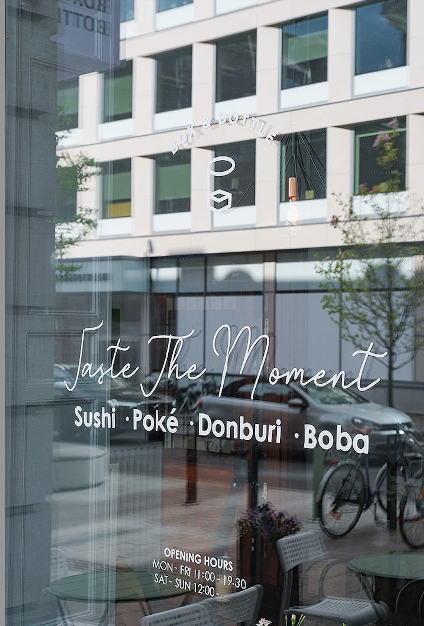 Restaurant window displaying 'Taste The Moment' and menu items: Sushi, Poké, Donburi, Boba, along with opening hours. Reflection of building and street outside.