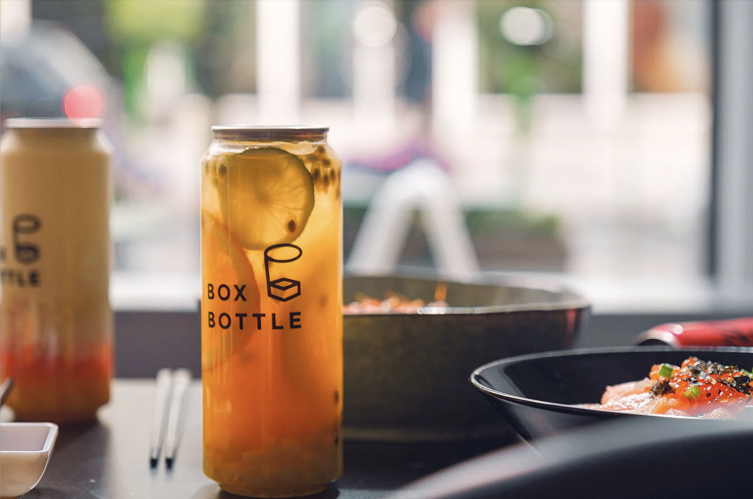 Beverage in a glass bottle with lemon slice, labeled "Box Bottle," next to a bowl of food on a table.