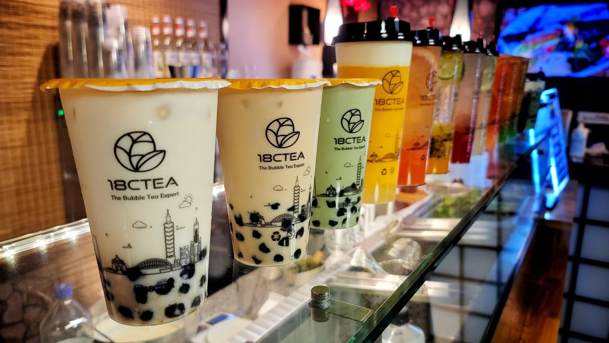 Row of bubble tea drinks with various colors and toppings, labeled with 18CTea, featuring tapioca pearls, on a glass counter in a shop.